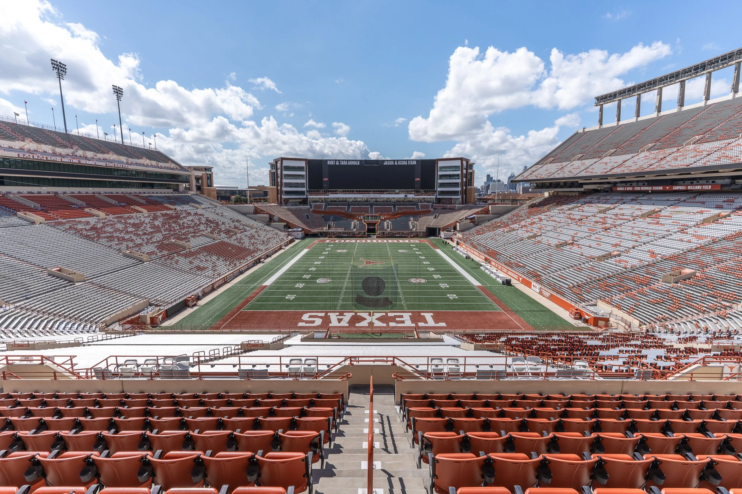 Empty football stadium with red and gray seats, a football field, and a large digital scoreboard under a partly cloudy sky.