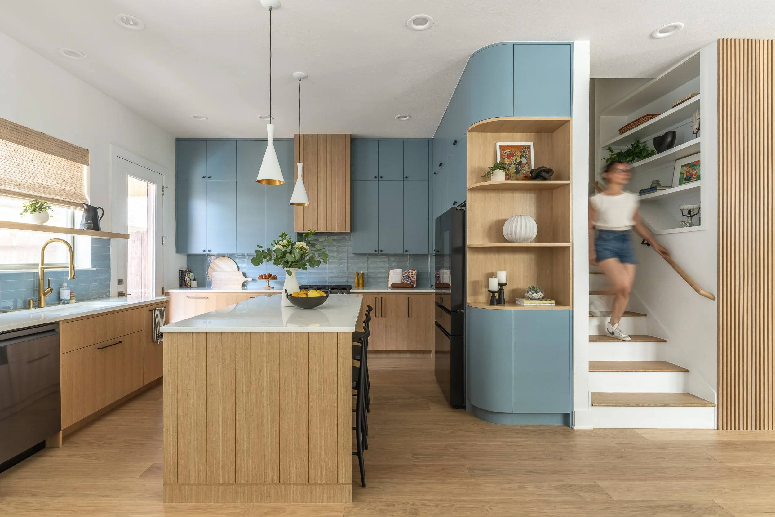 A modern open-concept kitchen with a large central island, blue upper and curved lower cabinets, and a woman walking up stairs on the right side.