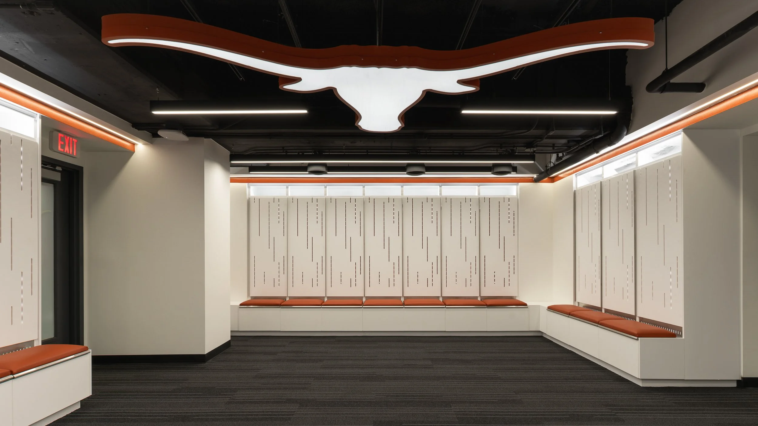 Modern waiting area with a long row of orange cushioned seats against white walls, a ceiling with a large decorative bull skull outline, and black ceiling with exposed pipes.