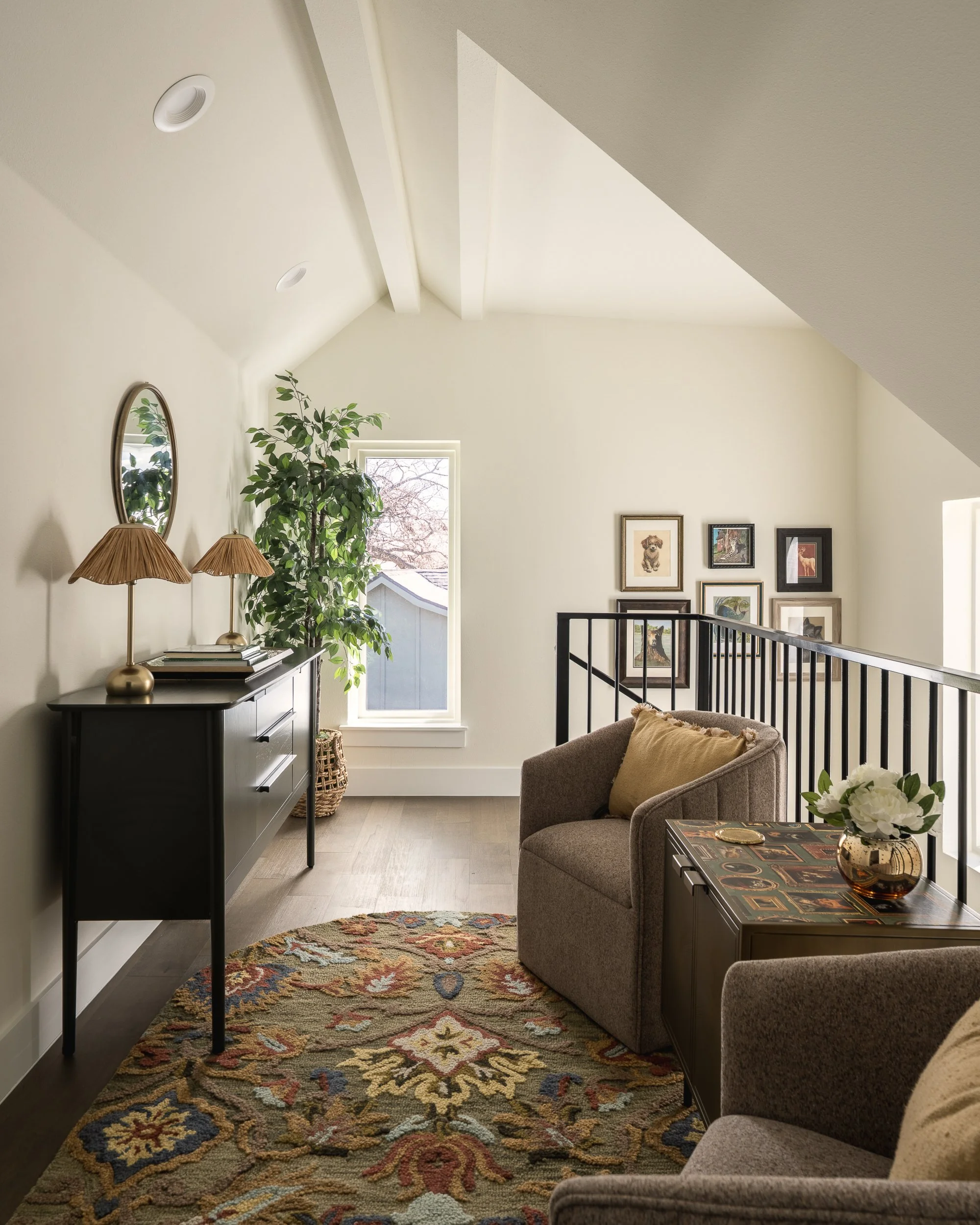 A cozy living room corner with a beige armchair and a side table with a vase of white flowers. A colorful patterned rug covers the wooden floor. A black console table against the wall has two gold-colored lamps, books, and a small mirror. A large gre