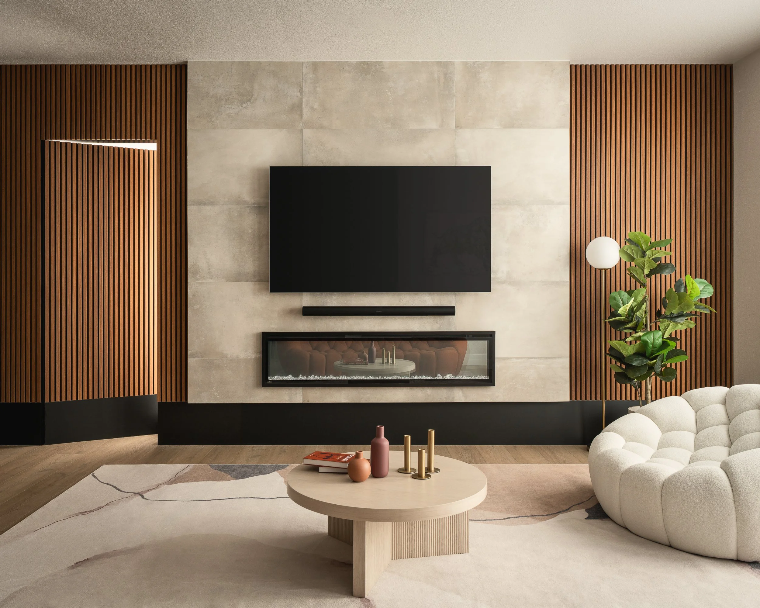 Modern living room with beige tiled fireplace, black accents, wooden slat walls, a round white coffee table with pink and gold decorative objects, a white curved sofa, a tall green plant, and a white spherical lamp.