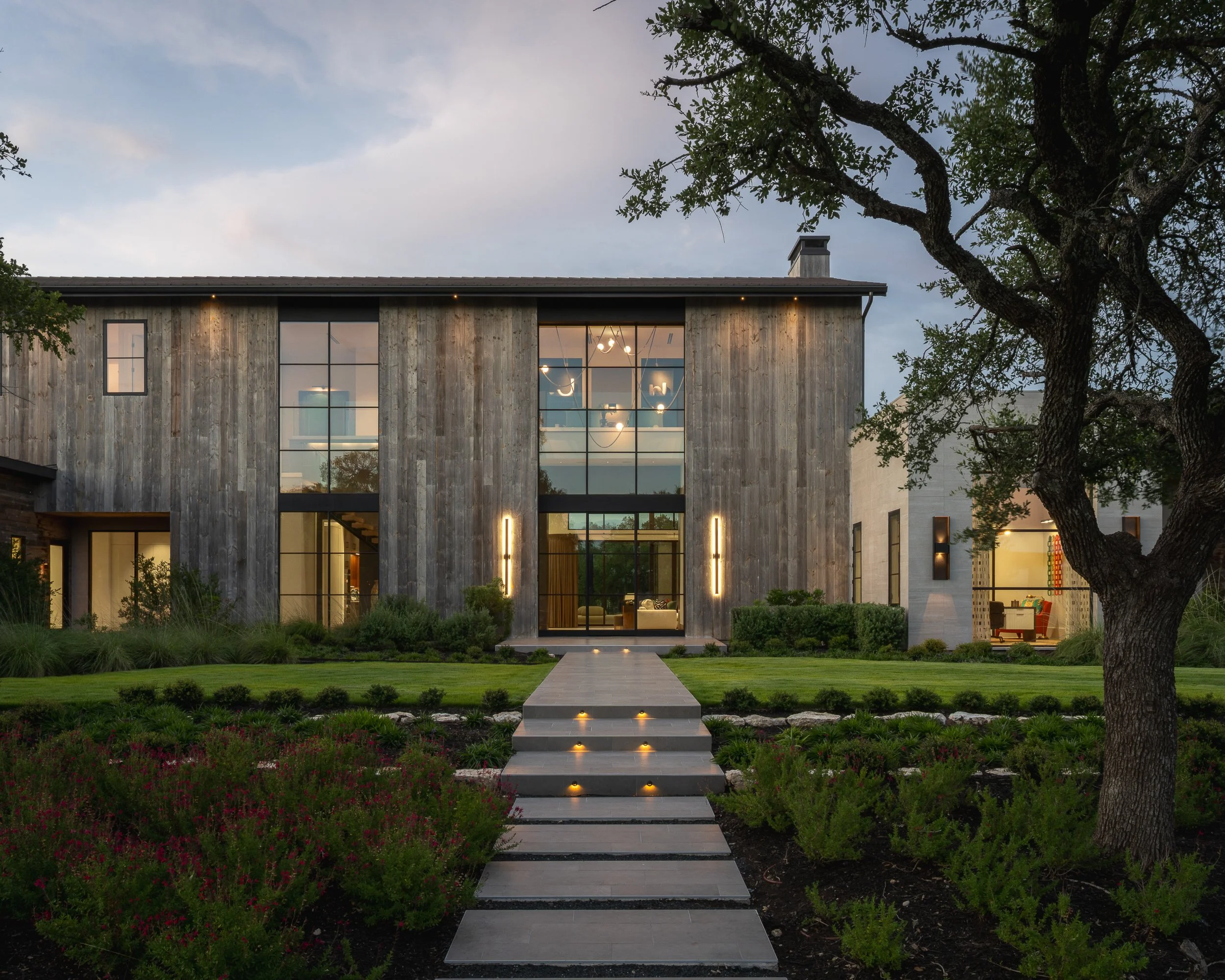 Modern house with large glass windows, surrounded by a well-maintained garden with a concrete pathway leading to the front entrance, illuminated by small ground lights at dusk, with a large tree in the foreground and a partly cloudy sky overhead.