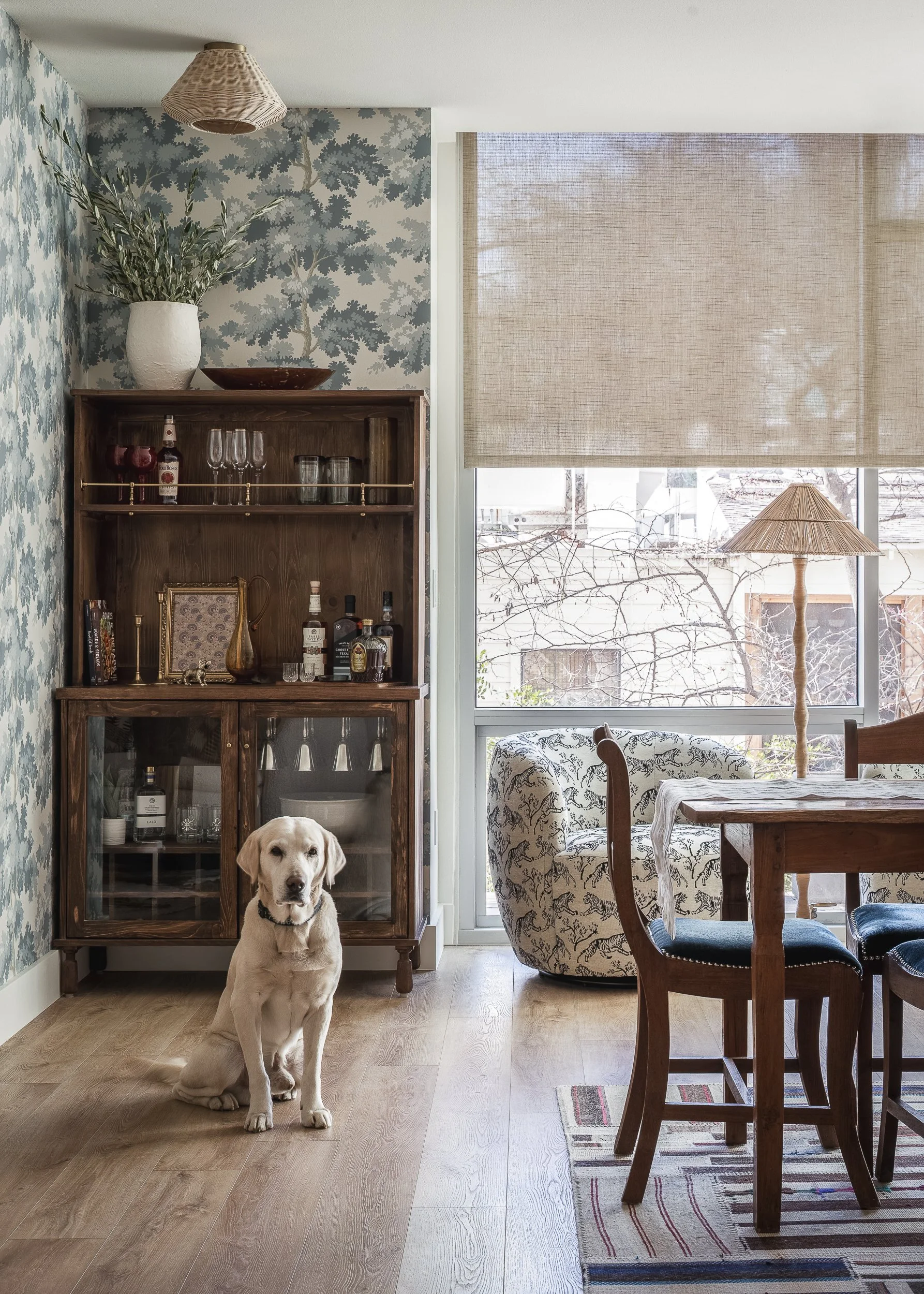 A Labrador puppy sitting on a wooden floor in a dining room next to a wooden cabinet with glass doors. The room has a large window with beige blinds, a patterned armchair at the corner, a wooden dining table with chairs, and a floor lamp with a woven