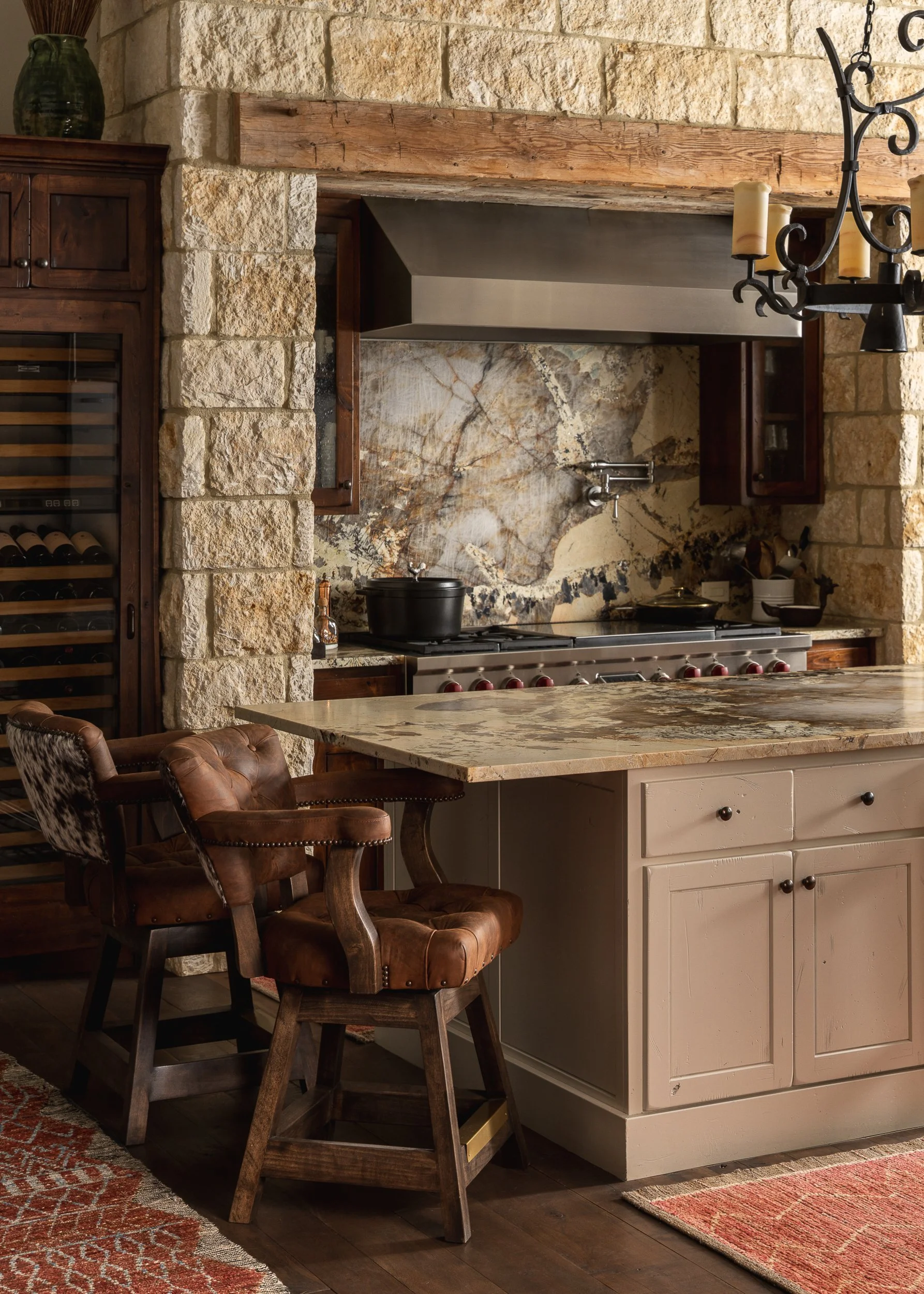 A cozy kitchen with a rustic stone wall and wooden accents. It features a white kitchen island with a variety of chairs at the counter, a wine cooler on the left, a large stone tile backsplash behind the stove, and a chandelier with candles hanging o