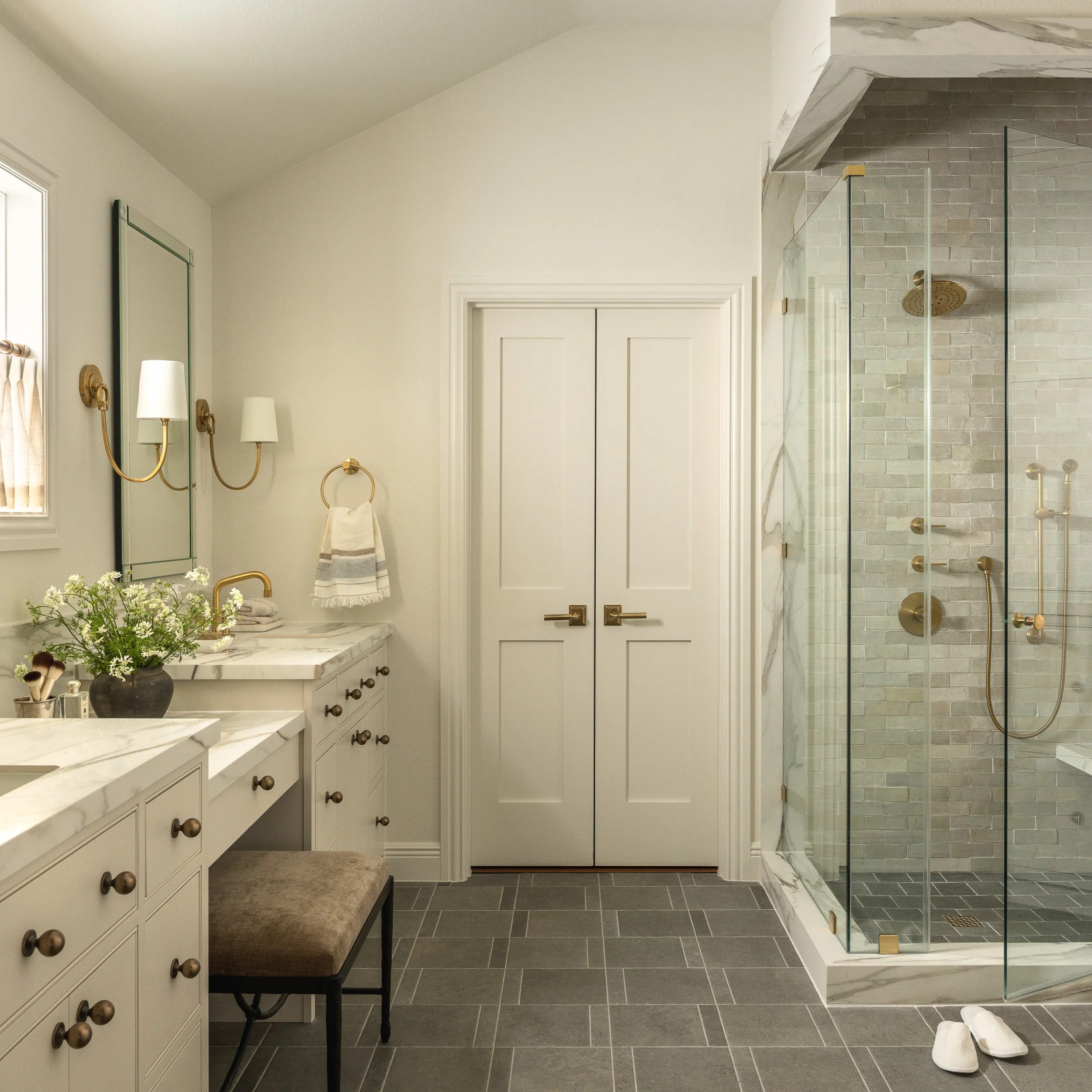 Modern bathroom with a marble countertop, a white double-door cabinet, a shower area with glass enclosure, gray brick tiles, and gold fixtures. There are white slippers on the gray tiled floor, and a towel with a striped towel on a bench.
