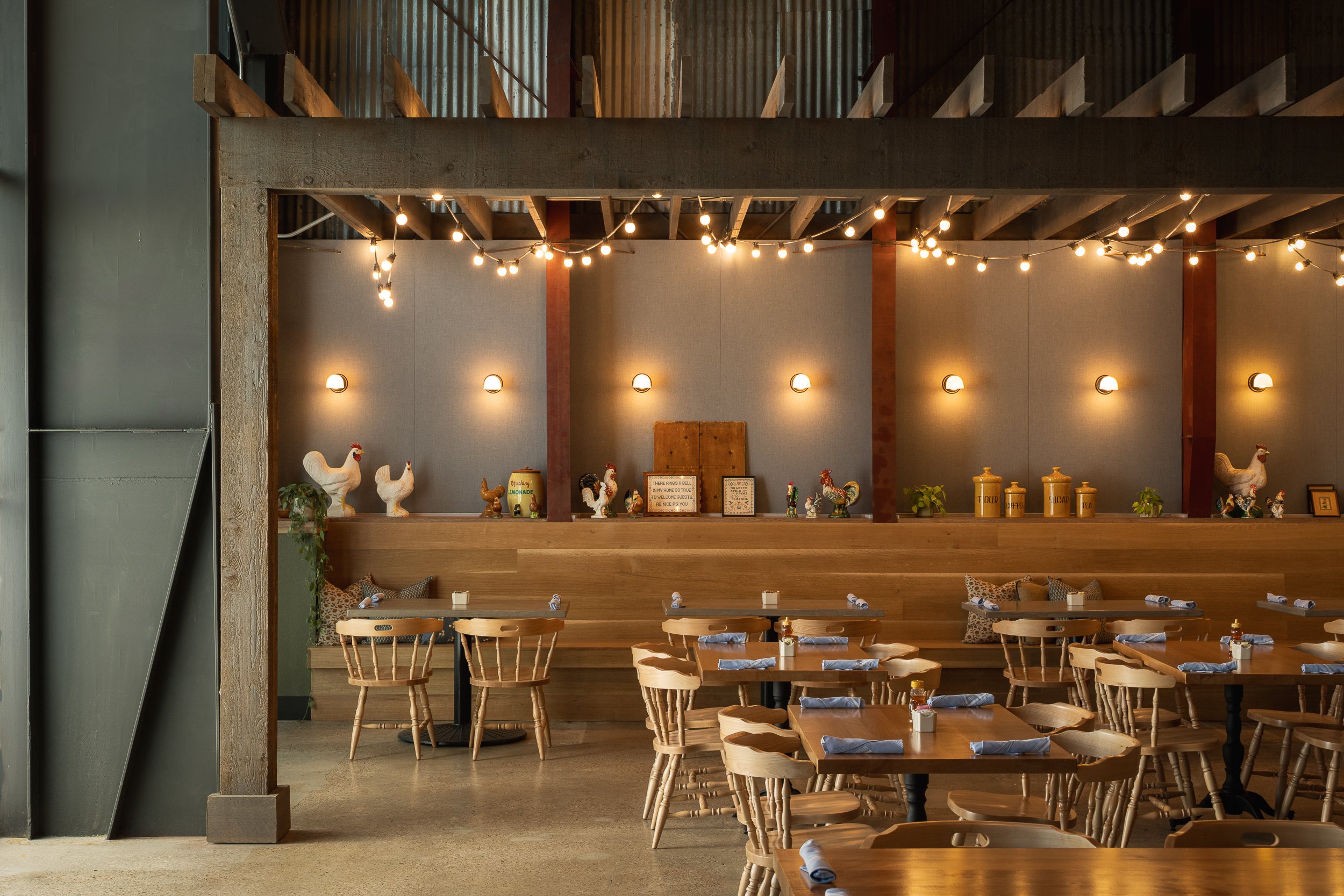 Interior of a cozy restaurant or cafe with wooden tables and chairs decorated with napkins. There are string lights hanging from the ceiling and ceramic rooster decorations on a high ledge. The wall is painted gray with small wall sconces providing a