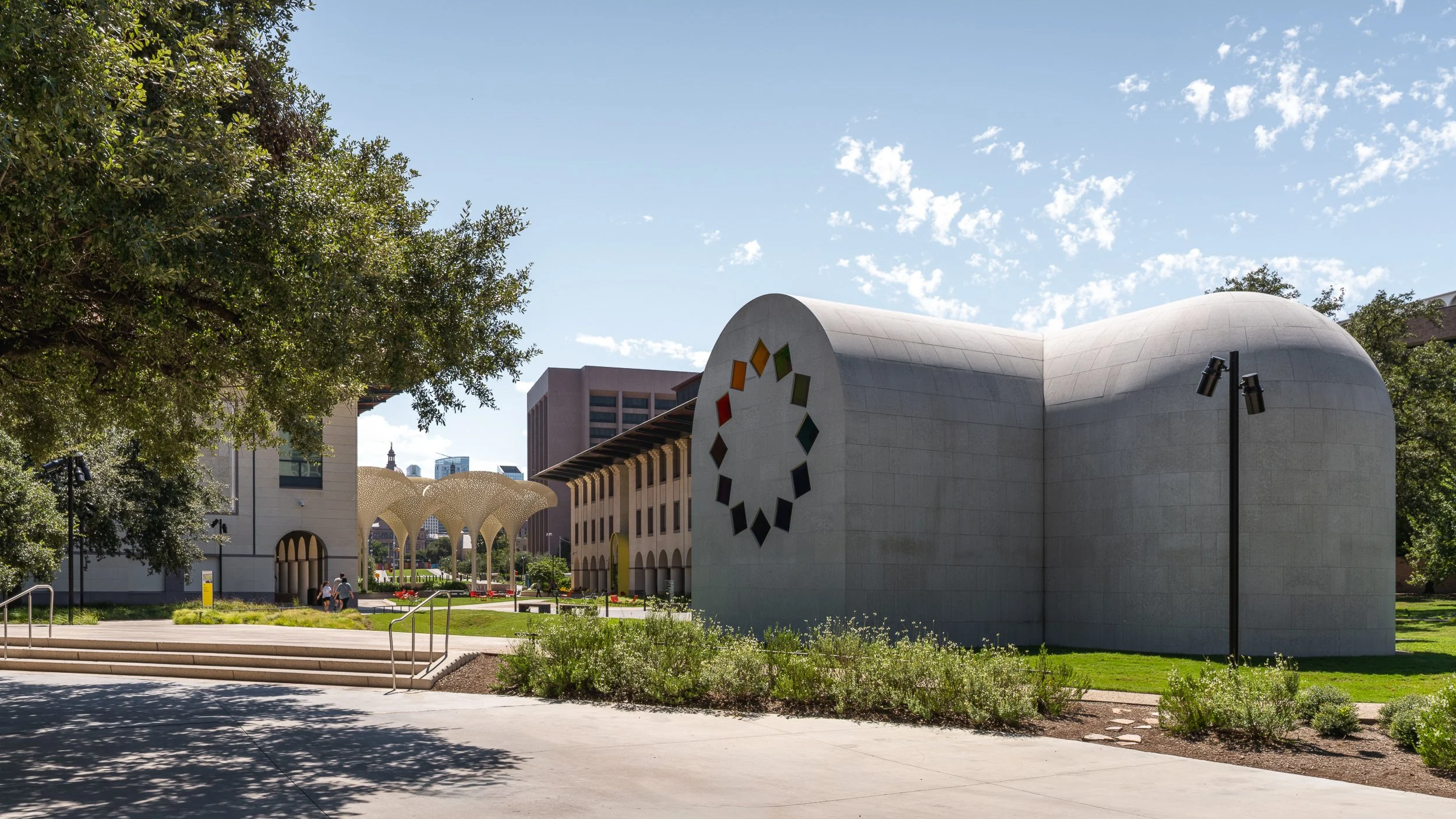 Modern building with colorful geometric design, surrounded by trees and open plaza with some people walking, under a blue sky with scattered clouds.