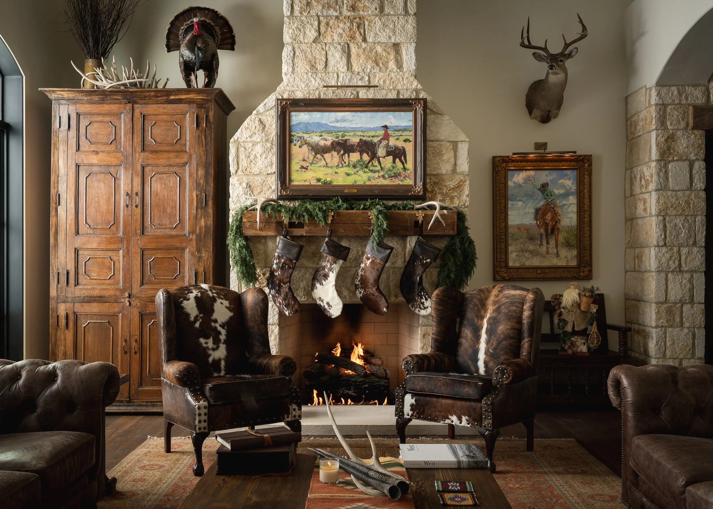 A cozy living room with a stone fireplace, decorated for the holidays. Above the fireplace, there is a landscape painting with horses and a cowboy. Mounted on the wall are a deer head and a turkey, along with some deer antlers. The room features leat