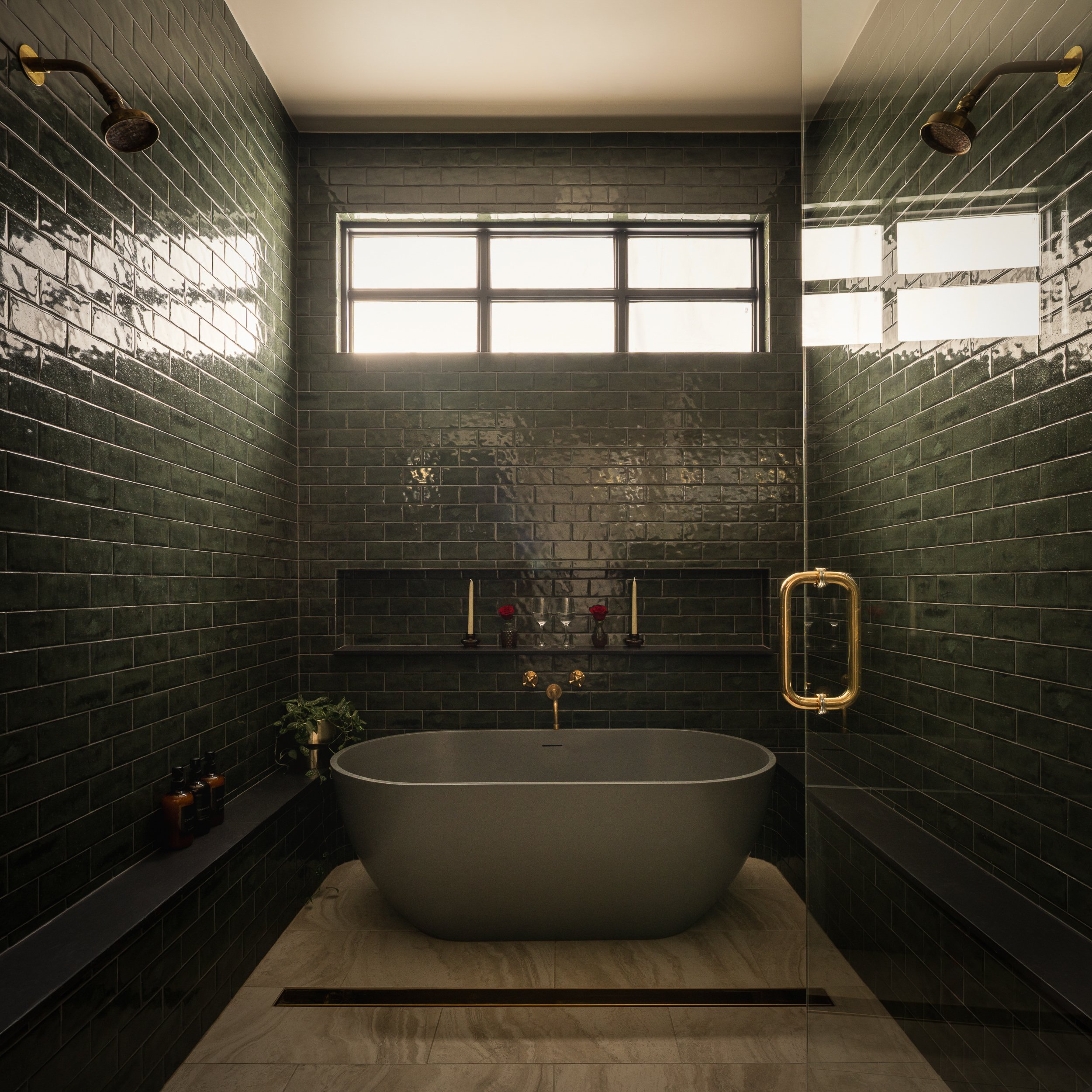 Modern bathroom with dark green tiled walls, a white freestanding bathtub, large window, and brass fixtures.