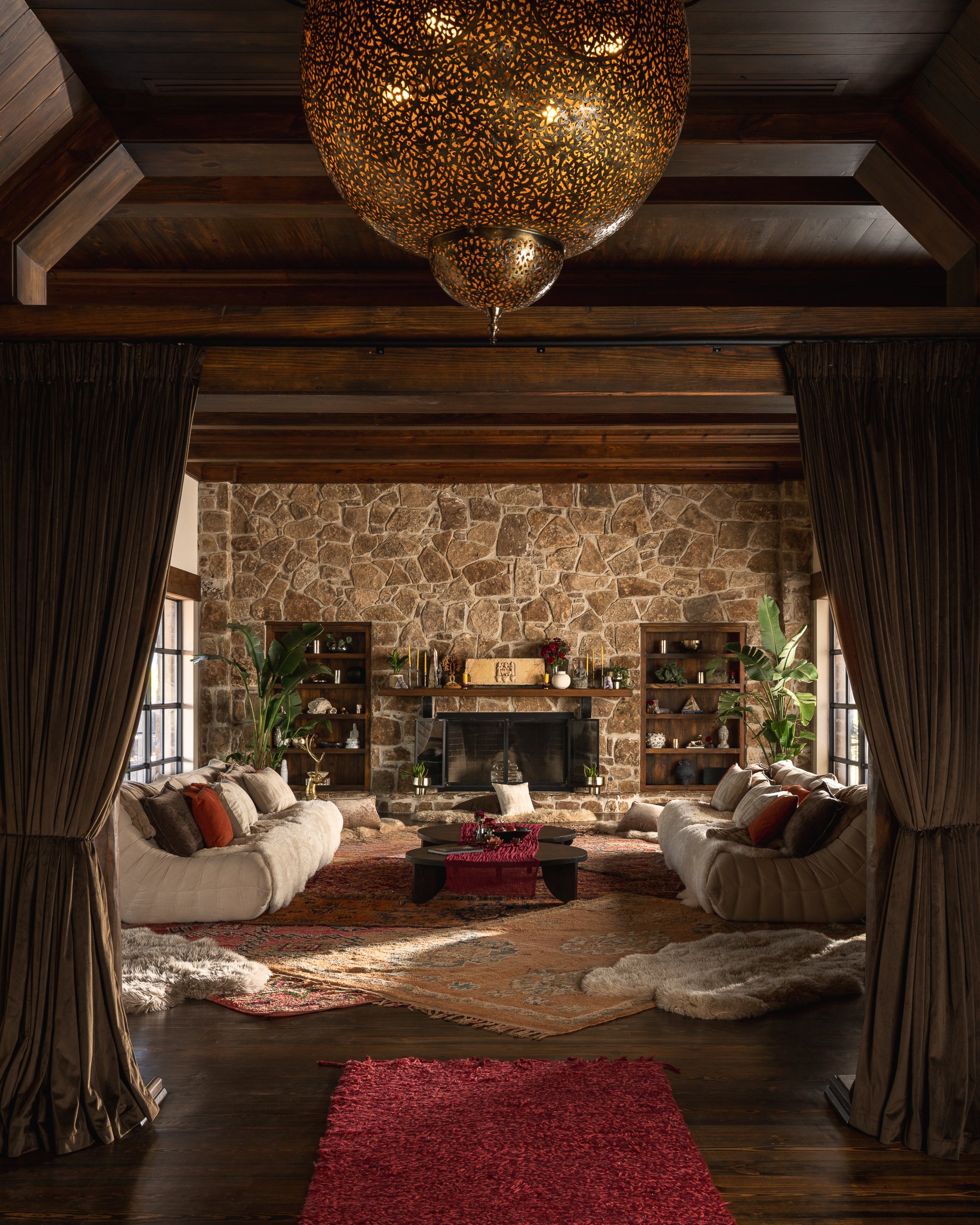 A cozy living room with a stone fireplace, wooden shelves, two beige sofas with pillows in front of large windows, an ornate pendant light hanging from a wooden ceiling, Persian rugs covering hardwood floors, and green plants on either side of the fi
