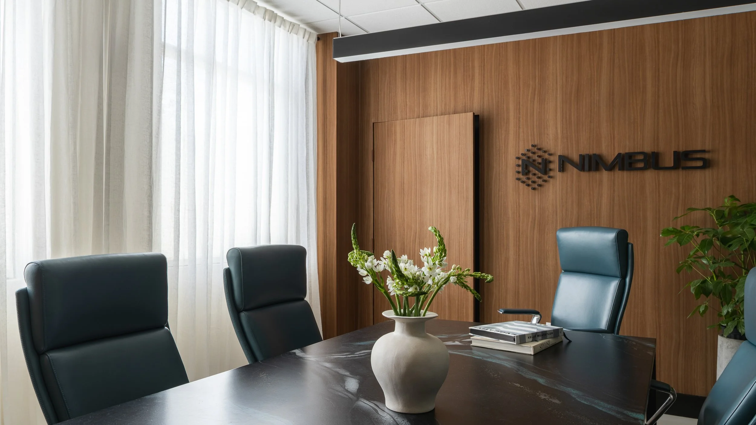Modern conference room with a wooden accent wall, a large table with a floral centerpiece, and four black leather chairs. There are large windows with white curtains and a potted plant in the corner. The wall features a sign that reads 'NIMBUS'.