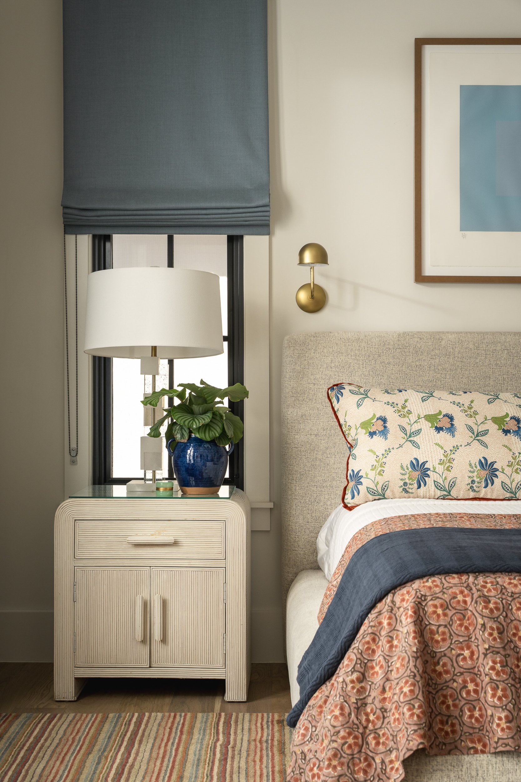 A bedroom corner with a beige upholstered bed, floral bedspread, a beige nightstand, a blue ceramic vase with a green plant, a white table lamp, a window with a blue Roman shade, a striped multicolored rug, a framed modern artwork, and a wall-mounted