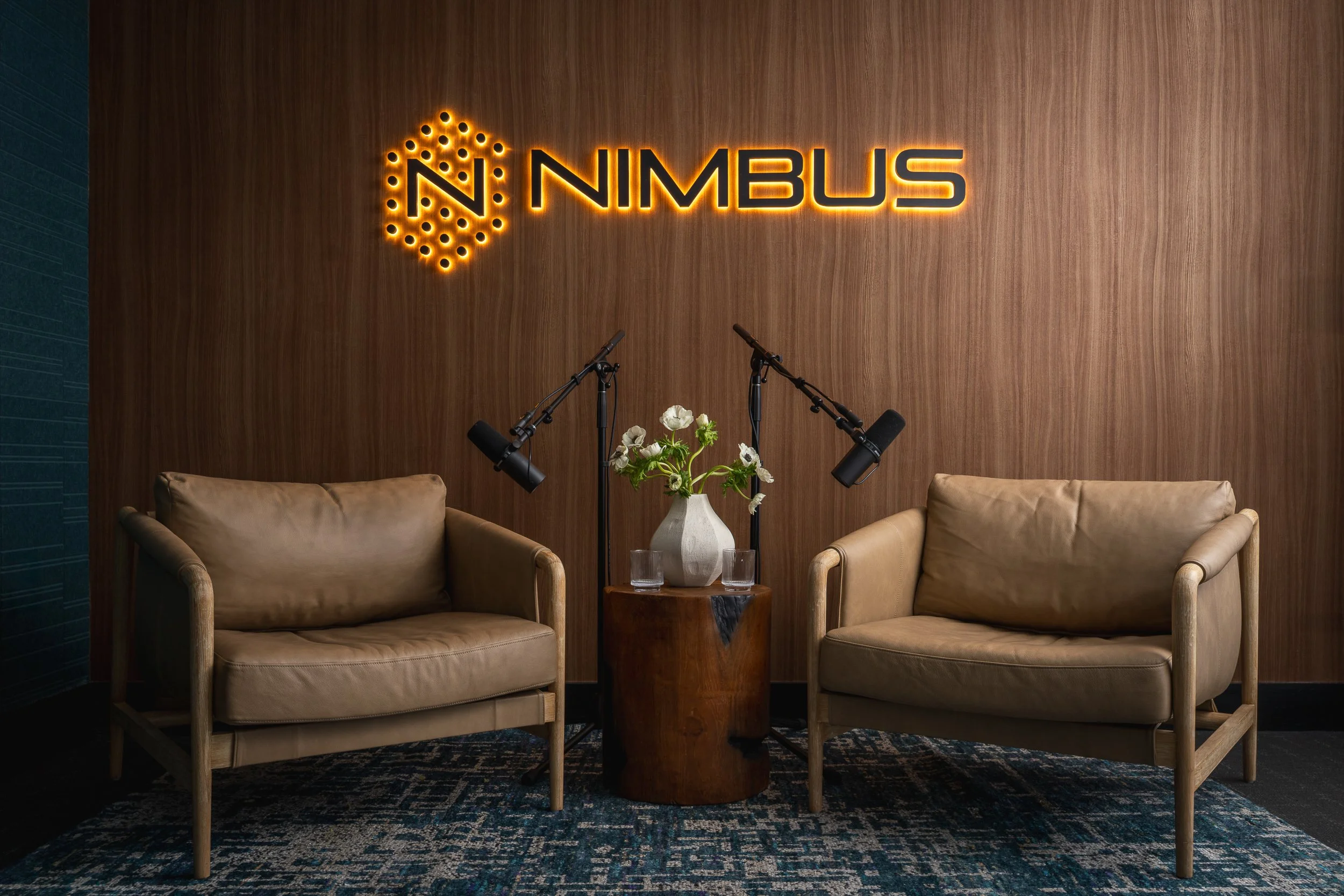 A room with a wooden wall and illuminated sign for NIMBUS. Two beige armchairs are on either side of a small wooden table with a white vase of flowers. Microphones are set up in front of the chairs, and a patterned blue and black carpet covers the fl