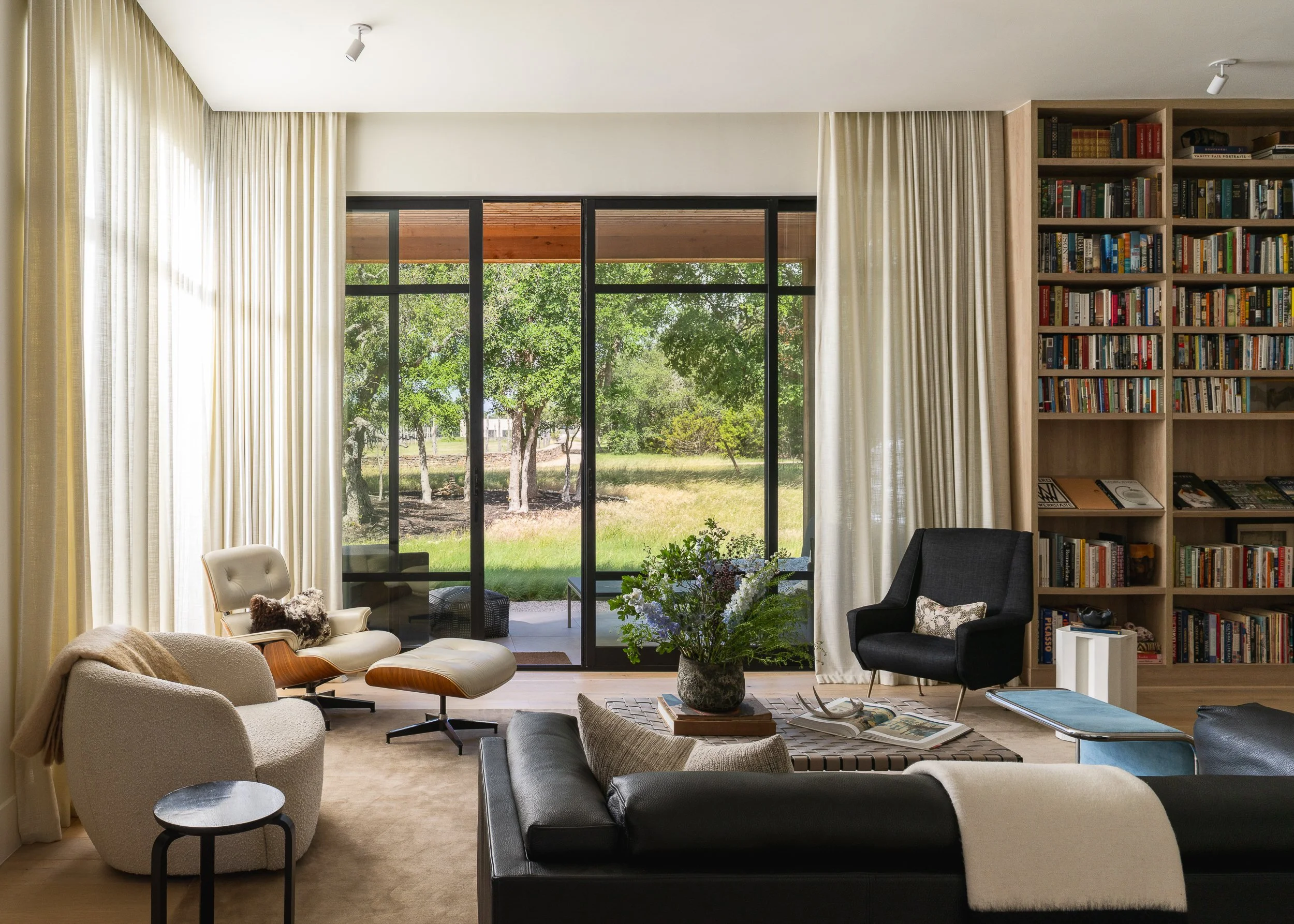 A living room with large glass sliding doors overlooking trees and greenery. The room has beige curtains, a black armchair, a white armchair with a wooden frame, a black rectangular coffee table with a floral arrangement, and a built-in bookshelf fil