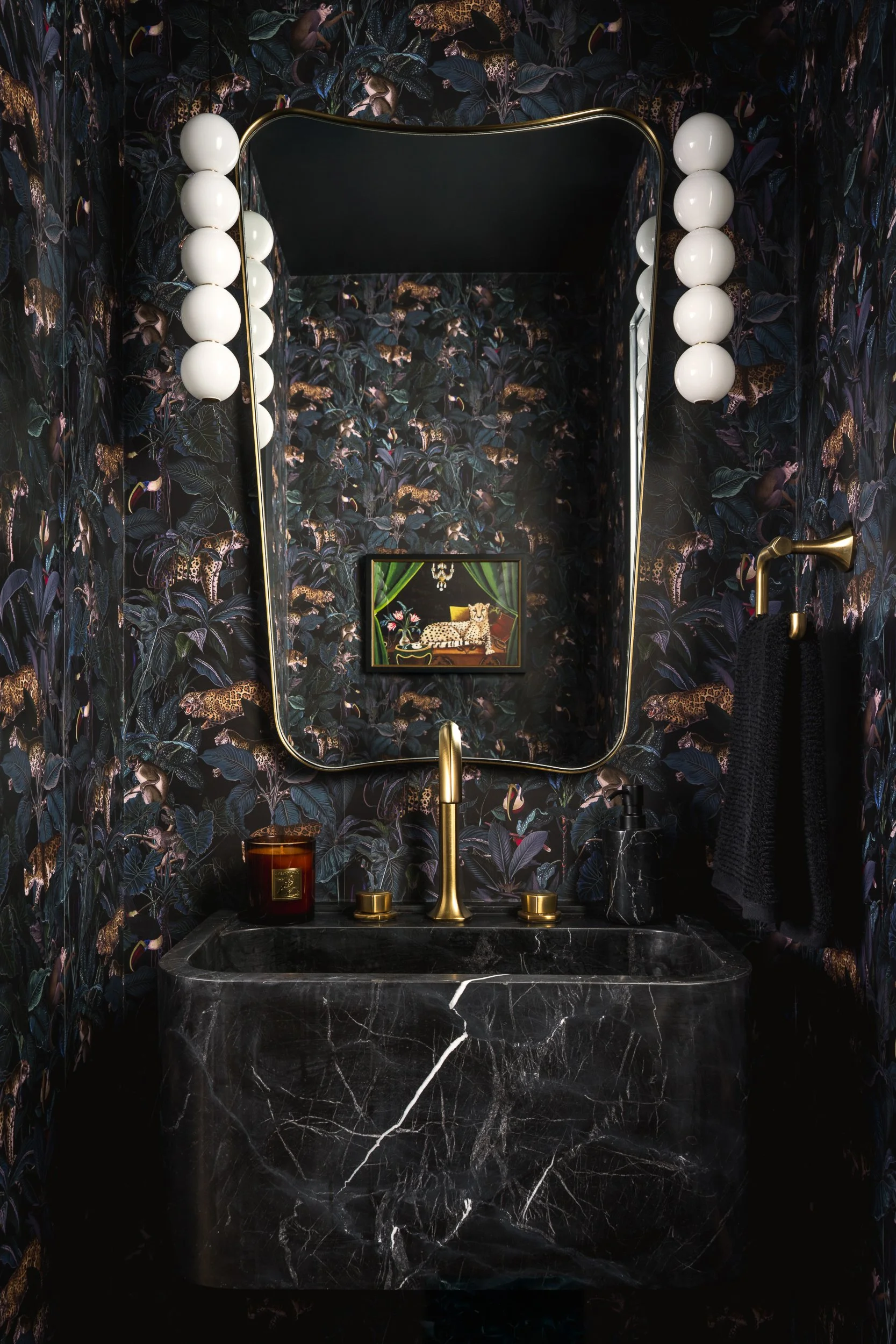 A bathroom sink area with black marble countertop, gold faucet, and black sink. The wall has dark jungle-themed wallpaper with animal patterns. Above the sink is a gold-framed mirror with round white bulbs on each side. A small framed picture of a ch