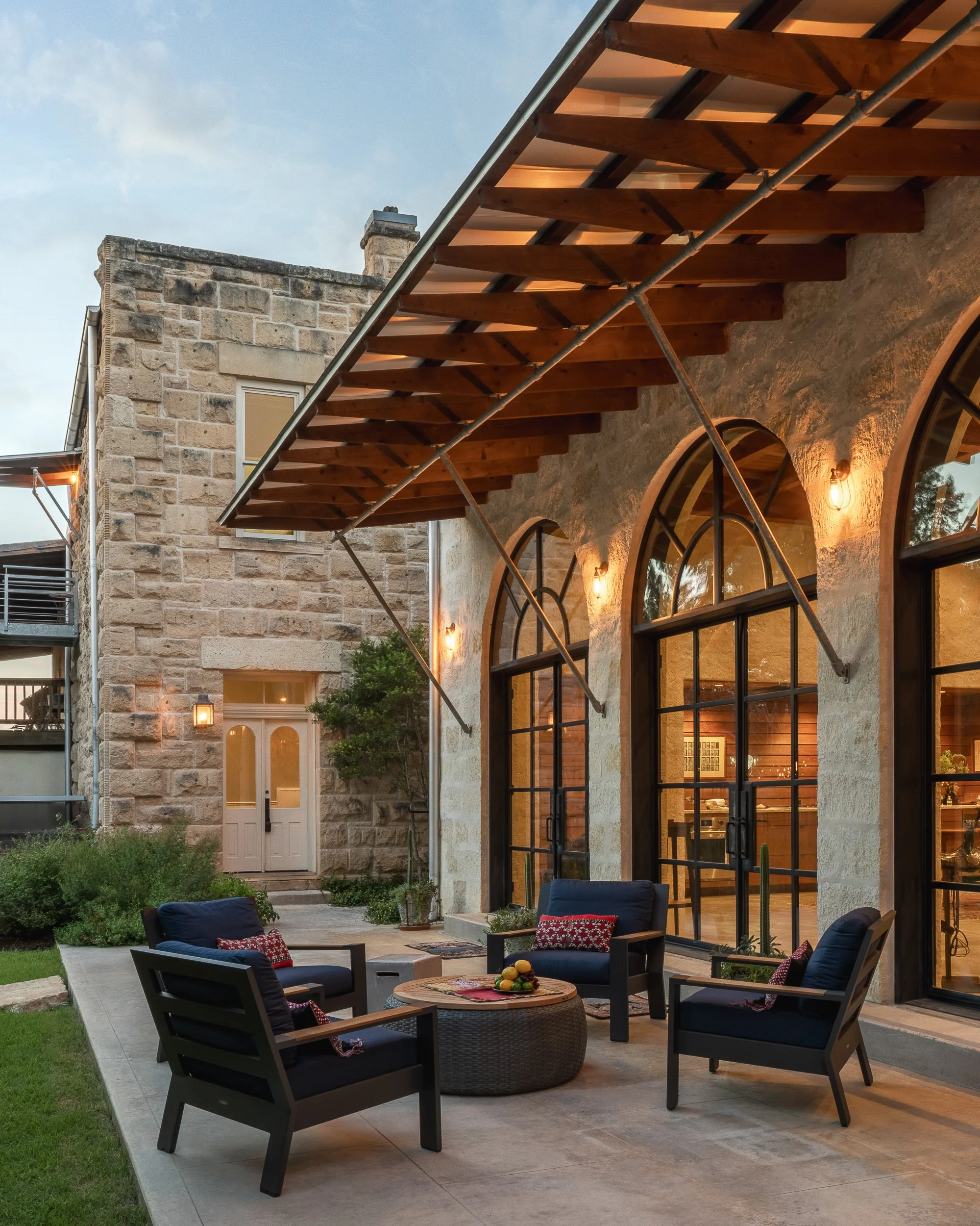 Outdoor patio furniture with dark blue chairs, a round table with a fruit basket, and a stone building with large arched windows and warm lighting, evening setting.