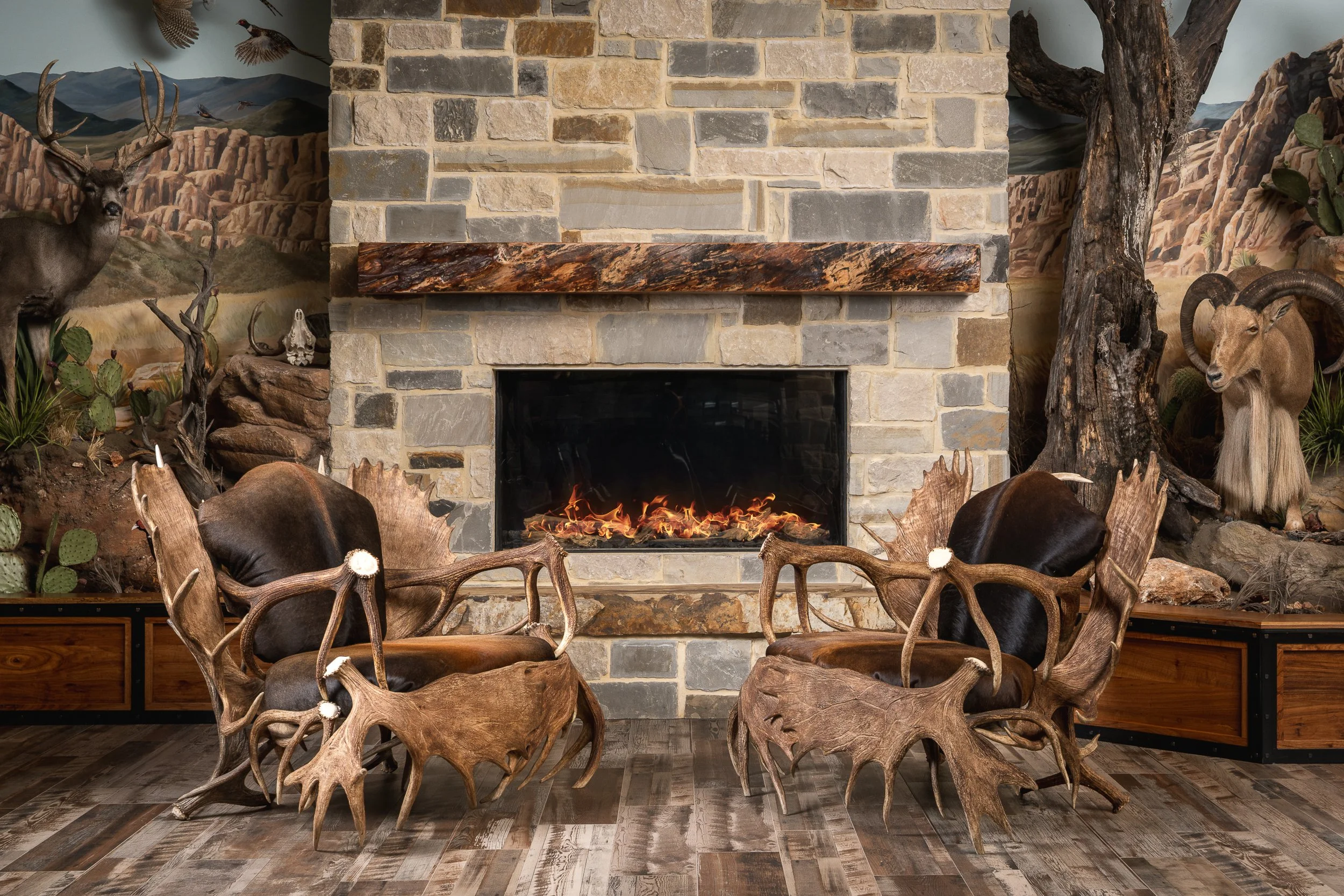 A rustic indoor living room featuring a stone fireplace with a wooden mantel. The room is decorated with taxidermy of deer and mountain goats, cacti, and a painted mountain landscape mural on the wall. Unique wooden chairs with intricate, natural sha
