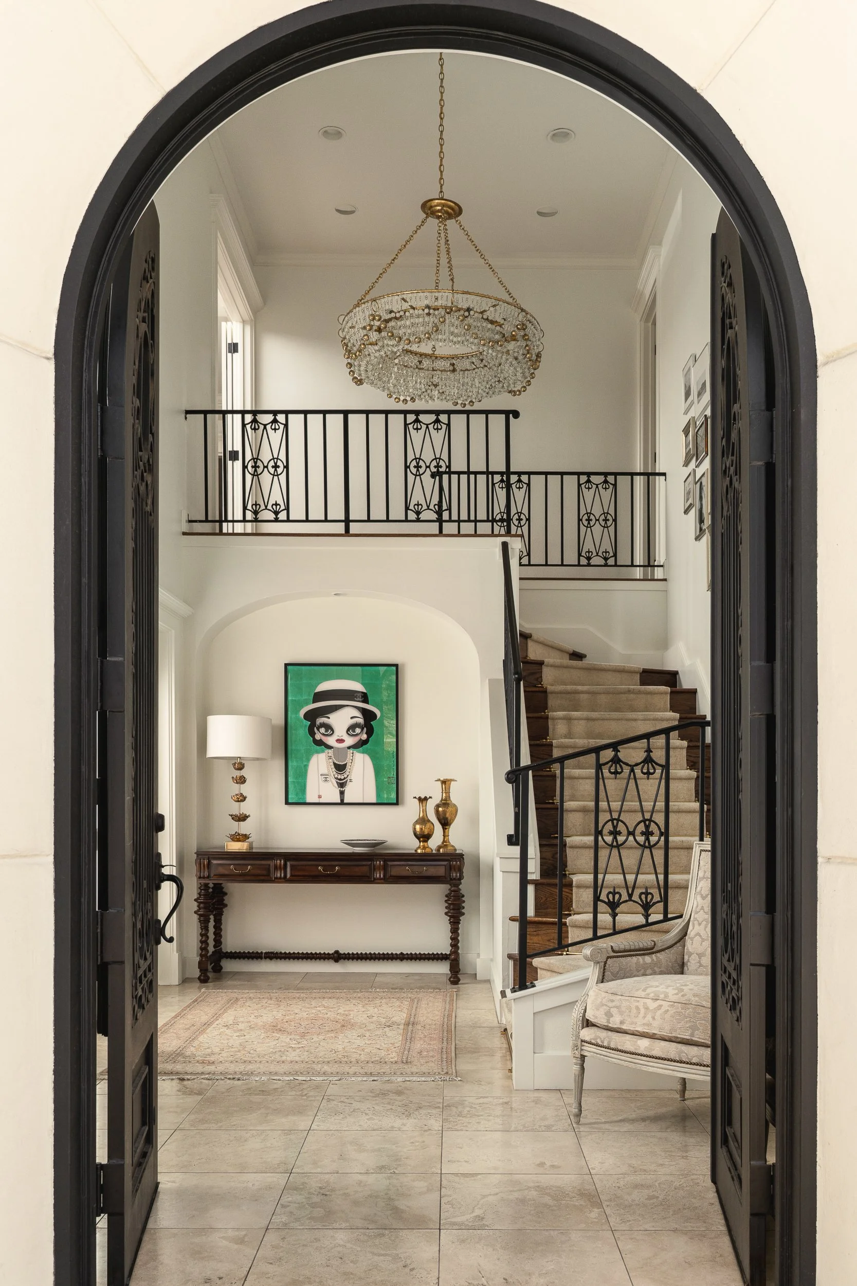 The image shows a grand, well-lit foyer seen from outside through an arched doorway with black iron gates. Inside, there is a staircase with dark wooden steps and black wrought iron railings, leading to an upper level. A large crystal chandelier hang