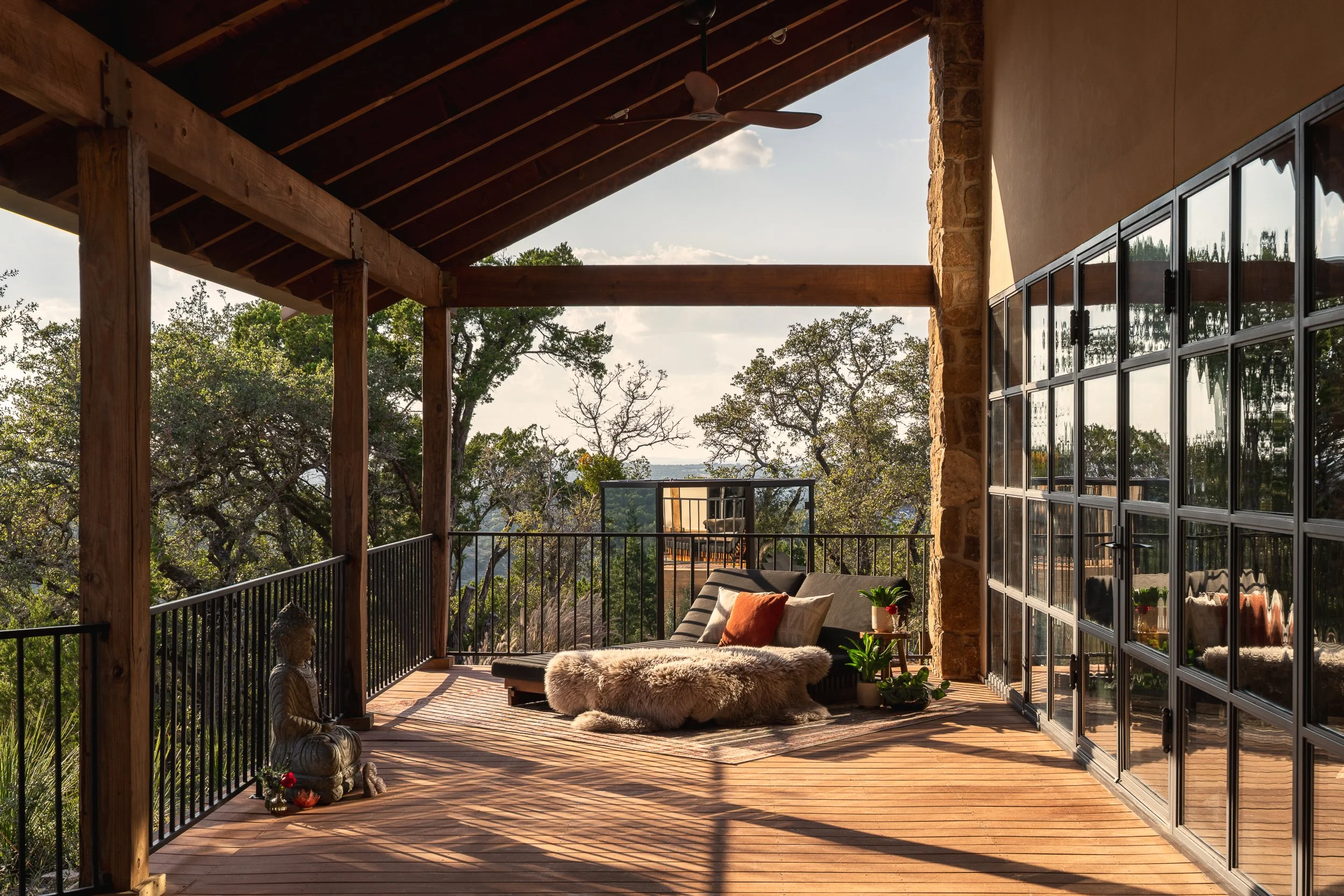 A spacious outdoor balcony with wooden flooring, black metal railing, and a ceiling fan. The balcony features a cozy seating area with a cushioned bench, fluffy throw blanket, and decorative pillows. It is partially enclosed with large glass doors, a