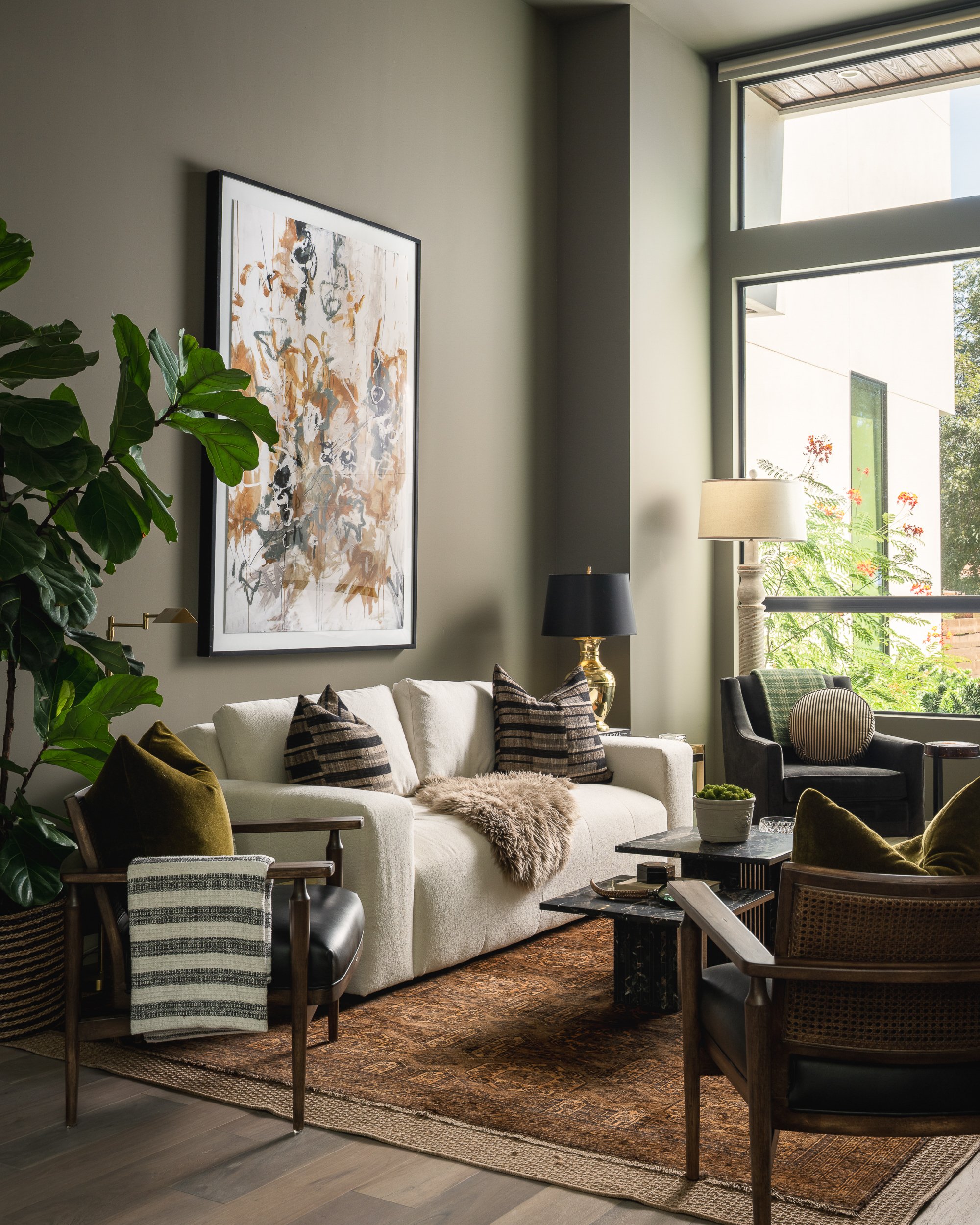 A cozy living room with a white couch decorated with striped pillows and a fluffy throw blanket. There are dark and patterned armchairs, a marble-top coffee table, and a vintage-style lamp. Large windows let in natural light, revealing greenery outsi