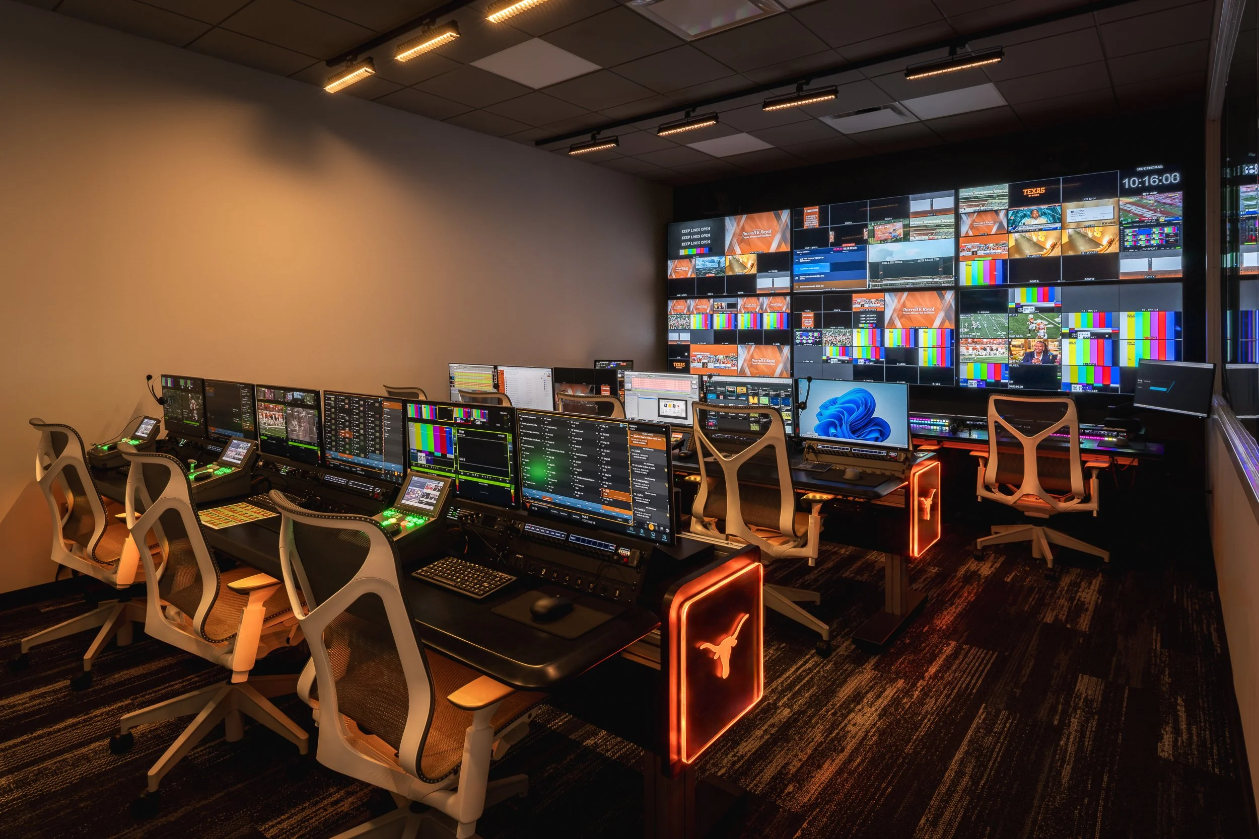 A television control room with multiple monitors displaying sports broadcasts, test patterns, and technical information. There are rows of desks with computer screens, control panels, and chairs, illuminated with warm lighting.