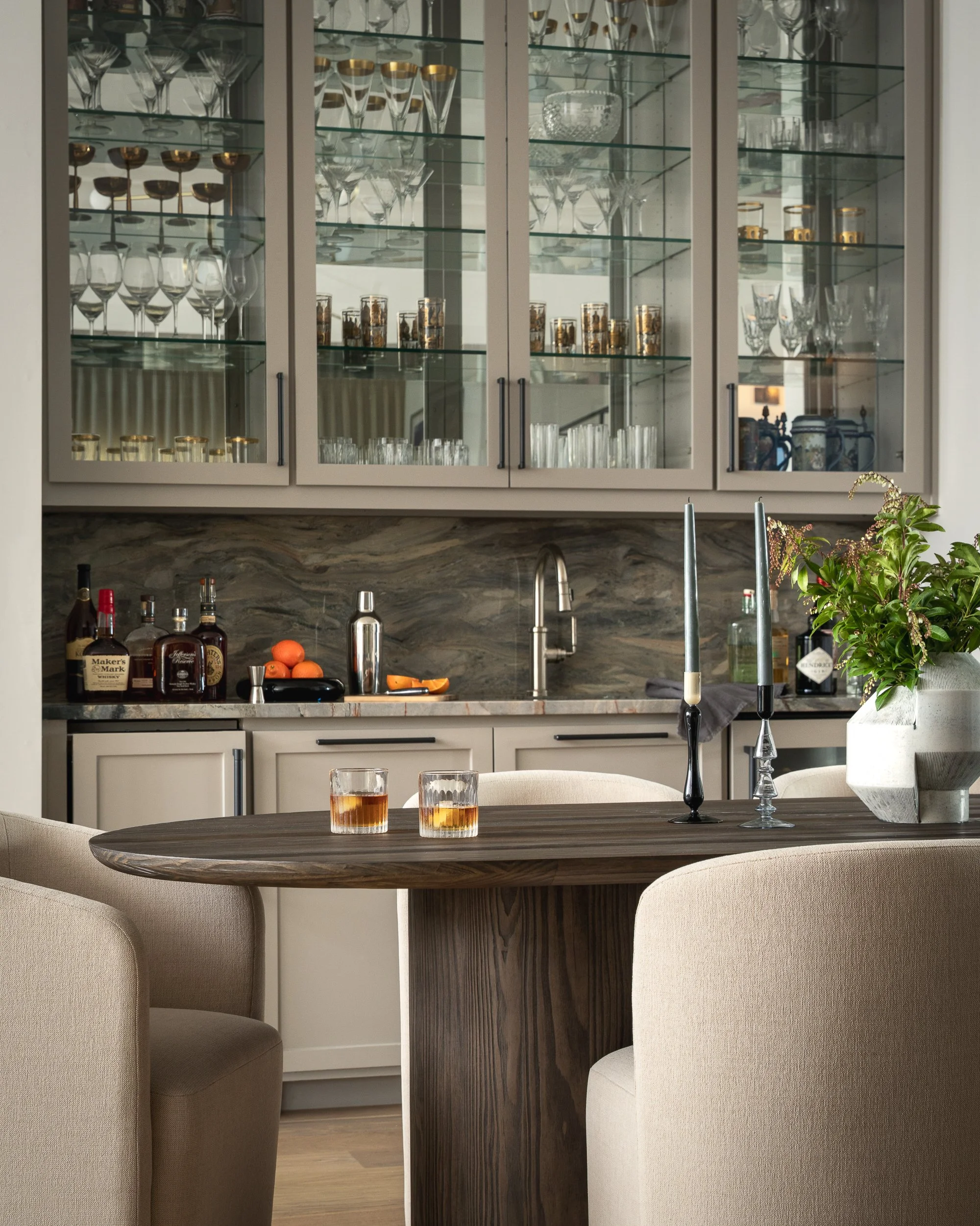 A kitchen with glass-front cabinets containing glassware, a marble countertop holding liquor bottles, oranges, and a knife, a round wooden table with drinks, padded chairs, a plant in a white pot, and a dark wood divider.
