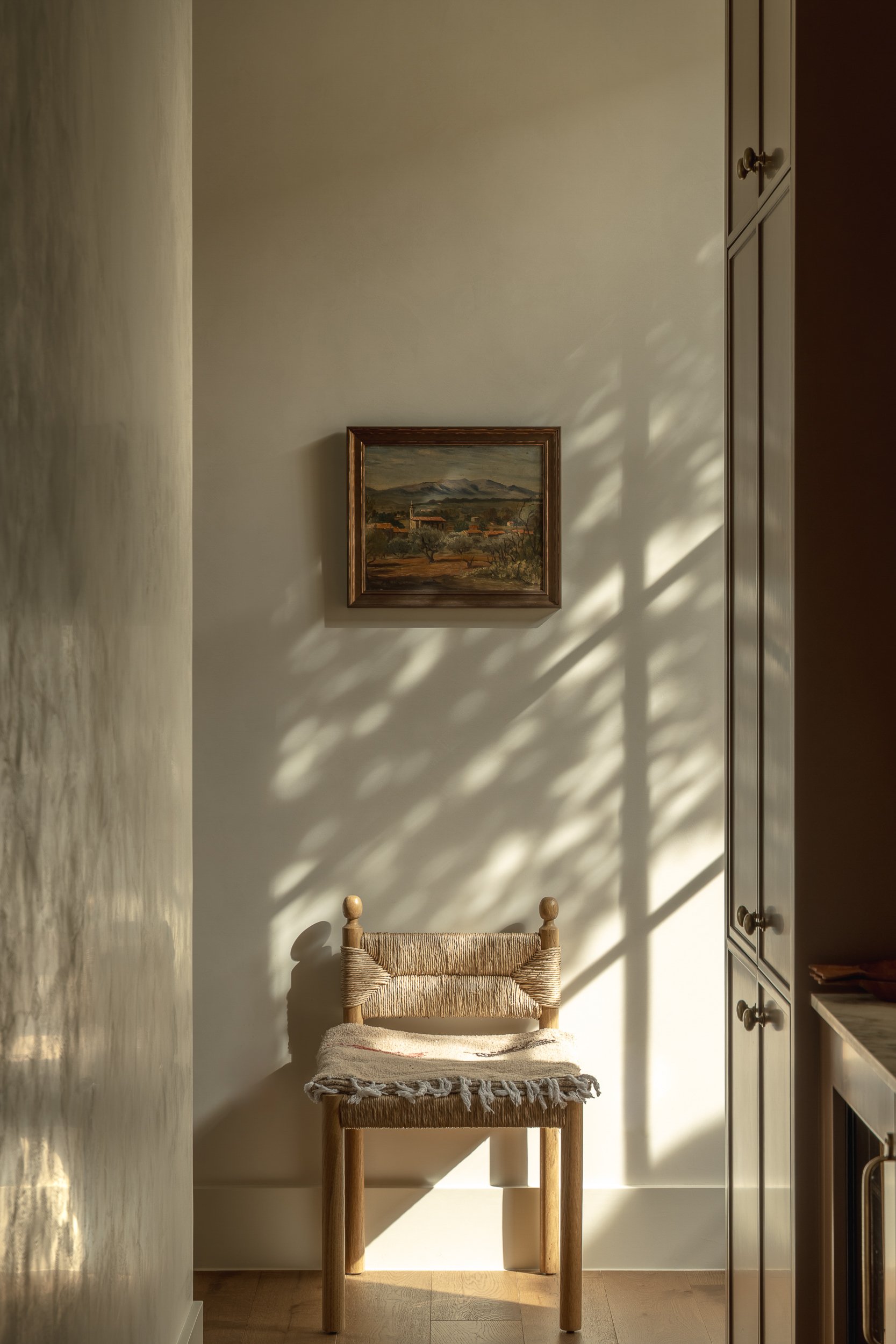 A small wooden chair with a woven seat and a blanket draped over it, positioned against a beige wall in a room illuminated by natural sunlight.
