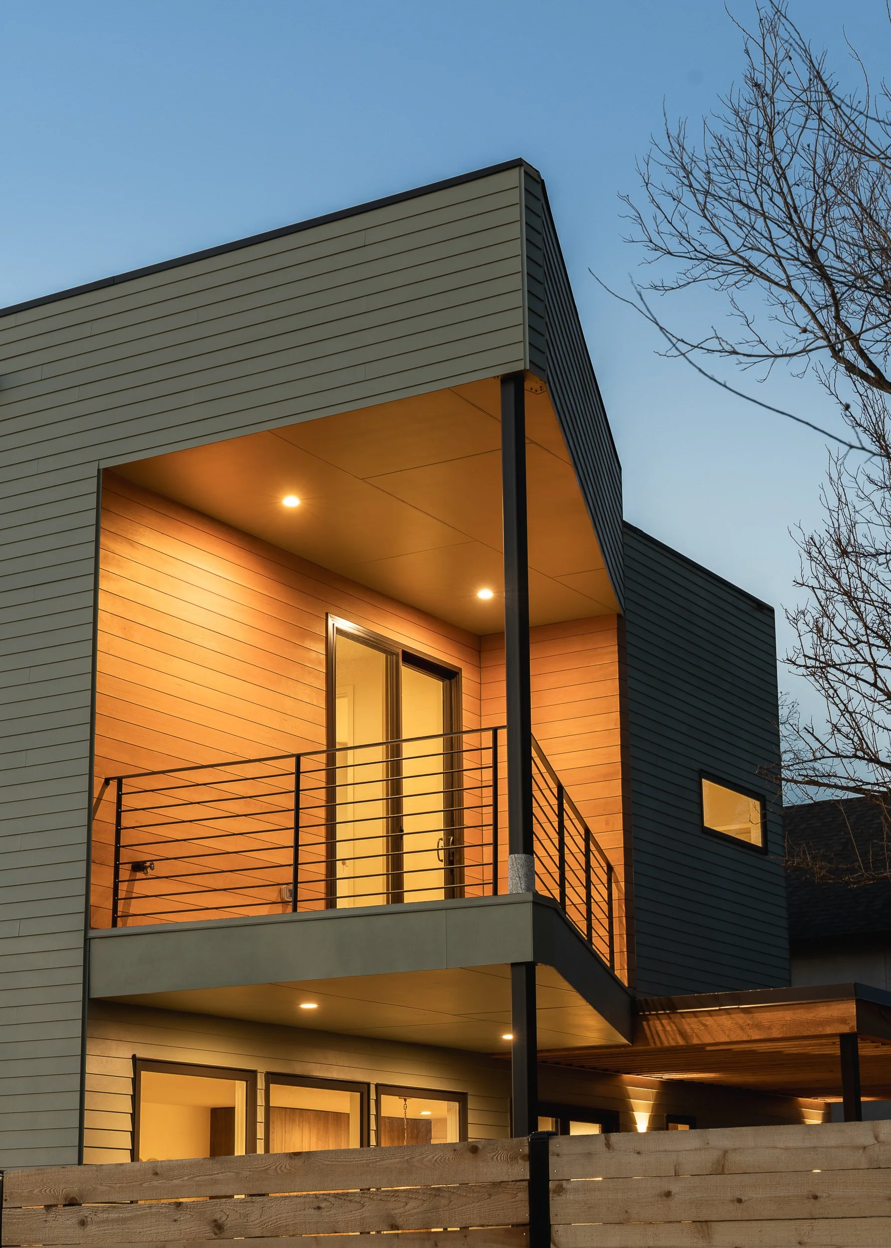 Modern multi-story residential building with lit windows and a balcony, surrounded by leafless trees, during dusk.