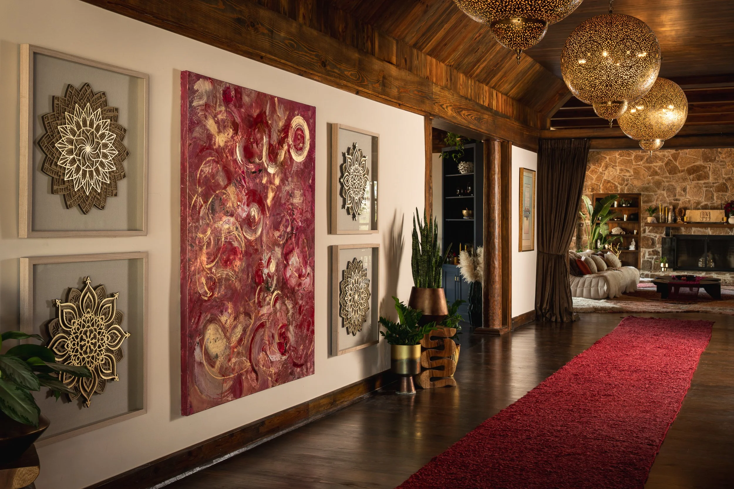 Living room with wall art, wooden ceiling, hanging lamps, fireplace, shelves, plants, and a red rug.
