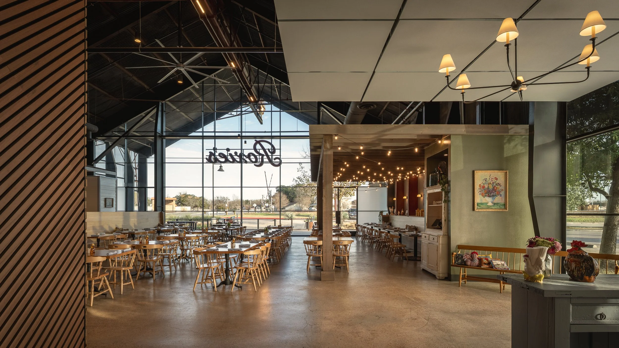 Interior of a modern restaurant with wooden tables and chairs, large windows with a sign that reads 'reataur', and decorative lighting including a chandelier and string lights.