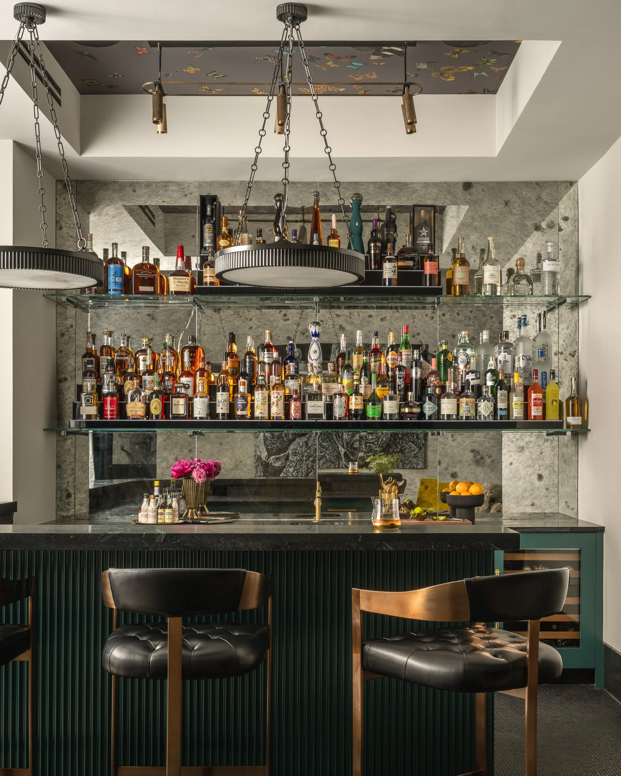 Bar area with two black leather chairs at a black marble counter, with a dark green paneling and a bar with varied liquor bottles in the background, and a vase with pink flowers on the counter.