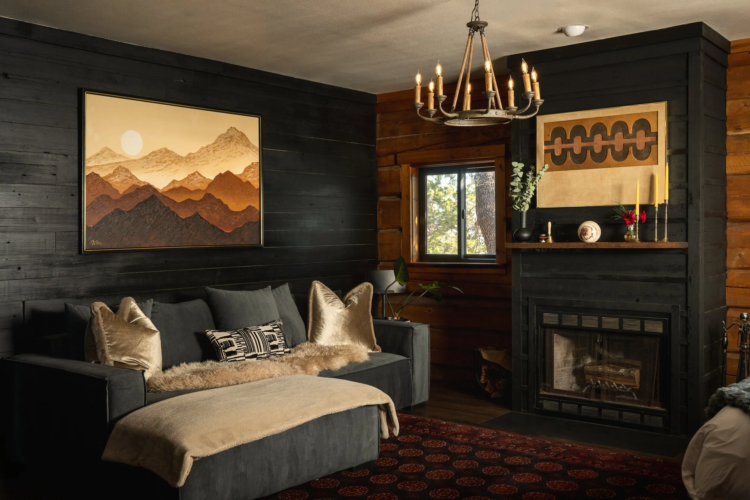 Living room with dark wood walls, a chandelier, a window showing trees outside, a mountain landscape painting, a black fireplace mantle with candles and decorations, and a gray sofa with beige and patterned pillows.