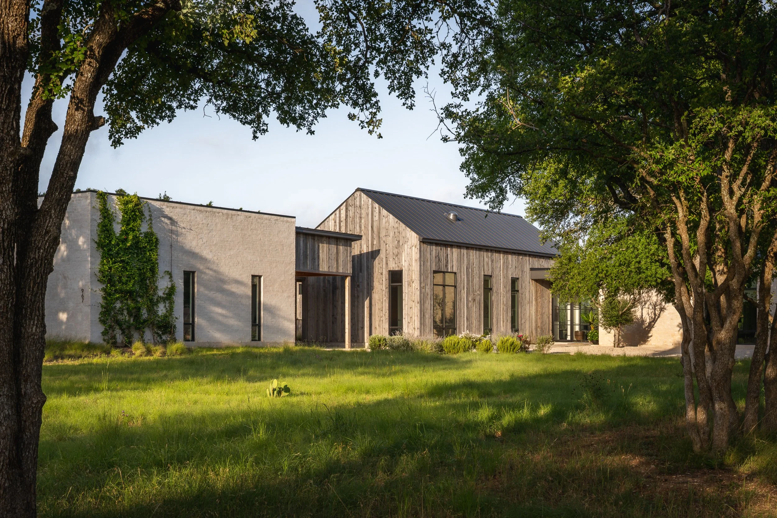 Modern house with a mix of white and wood exterior, surrounded by trees and greenery, under a clear blue sky.