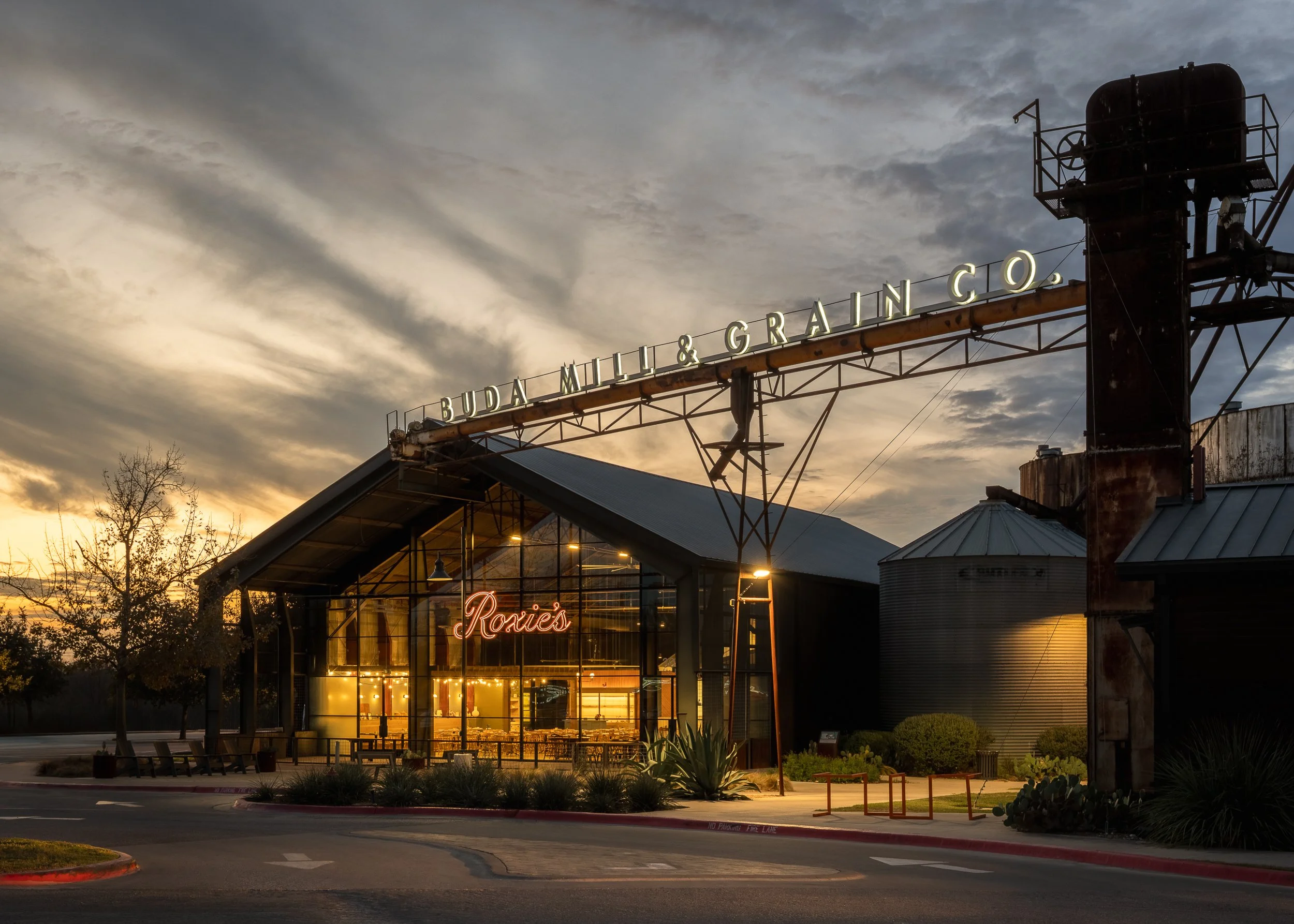 View of a restaurant or market named 'Roxie's' with a neon sign, large windows, and outdoor lighting, set against a cloudy sky at sunset. There are trees and landscaping around the building, and industrial structures like smokestacks are visible near