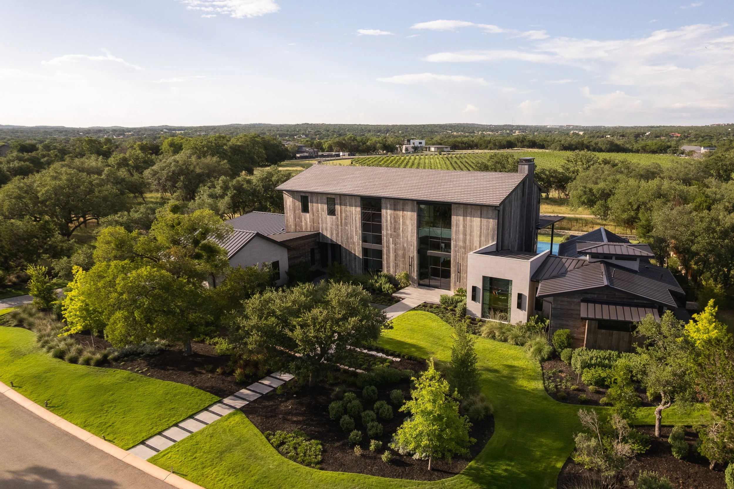 Aerial view of modern house with manicured lawn, trees, and surrounding countryside under a blue sky with clouds.