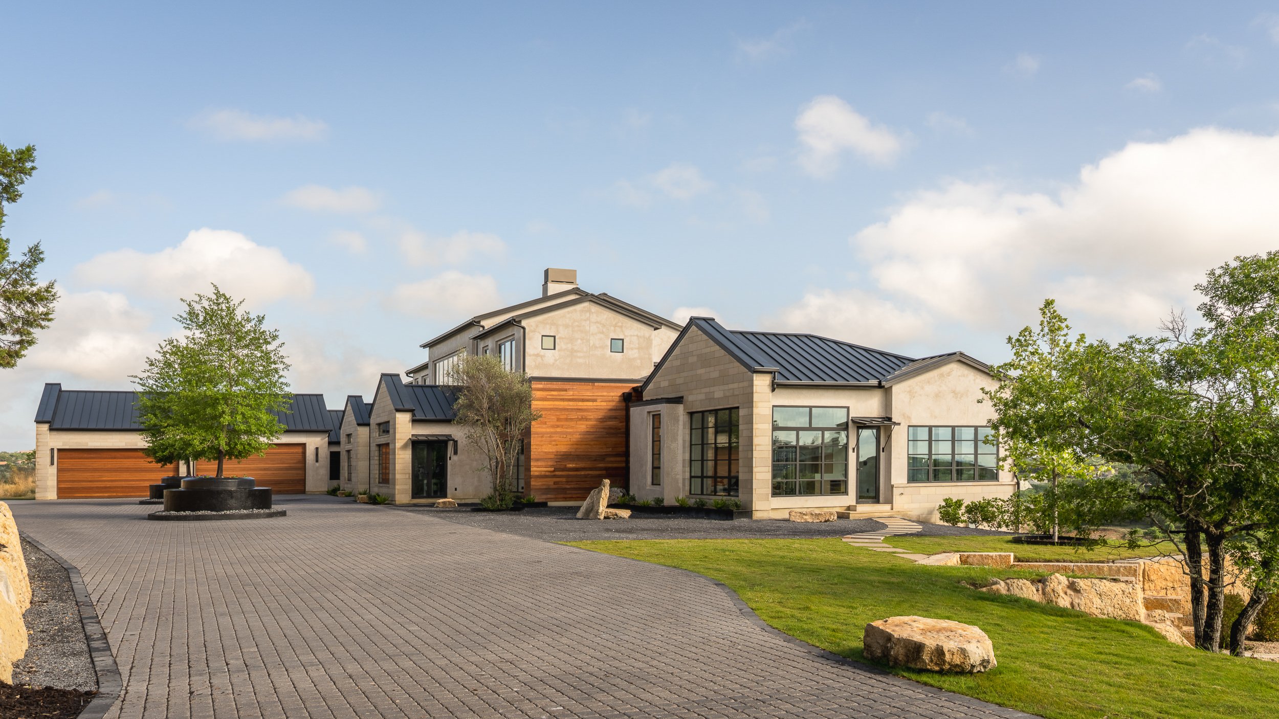 Modern house with large windows, a mix of stone and wood exterior, surrounded by trees, with a paved driveway and landscaped yard under a partly cloudy sky.
