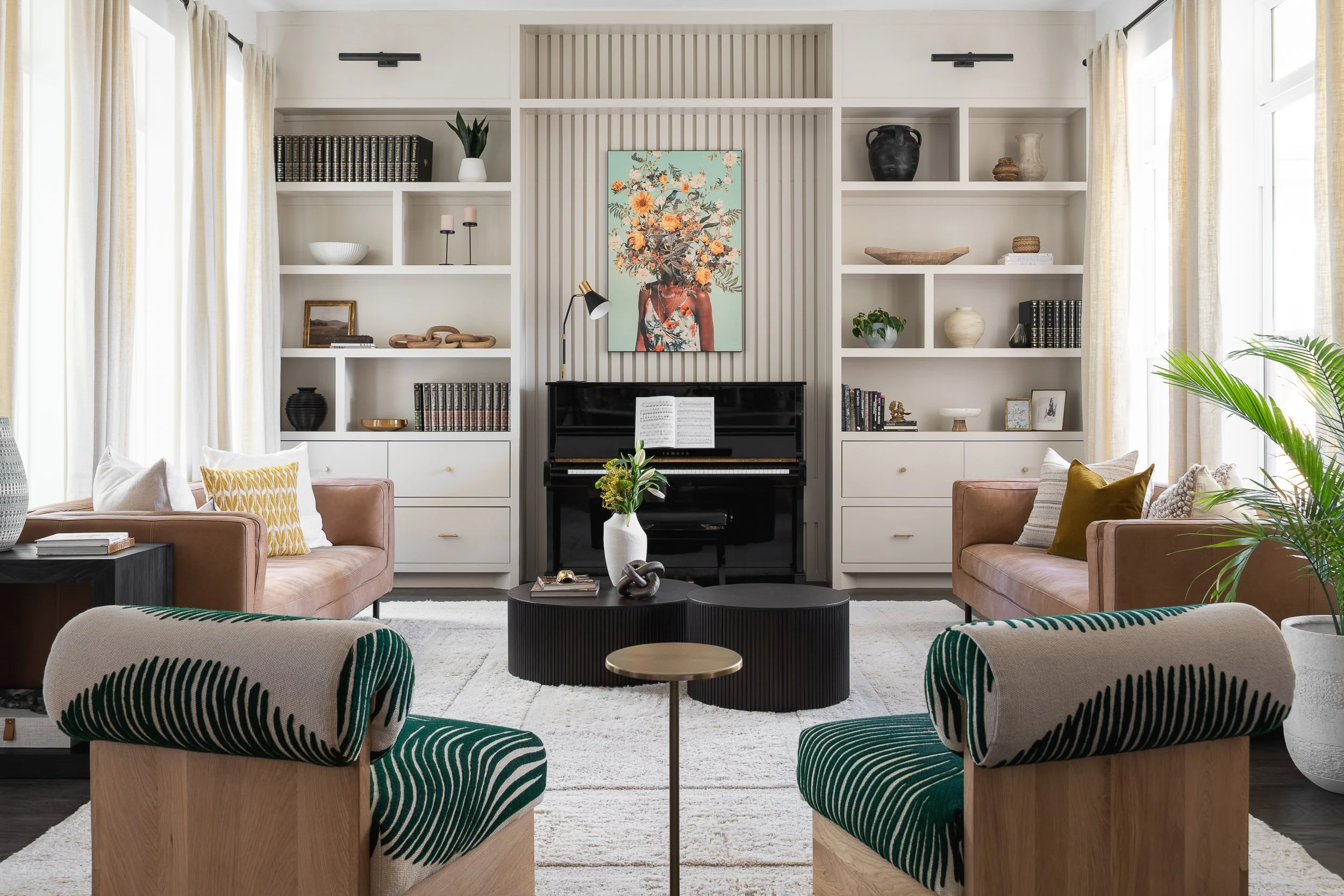 Living room with beige and green upholstered chairs, a black round coffee table, a white area rug, built-in white bookshelves with decorative items, a black upright piano with an open sheet of music, a colorful portrait of a woman with floral headdre