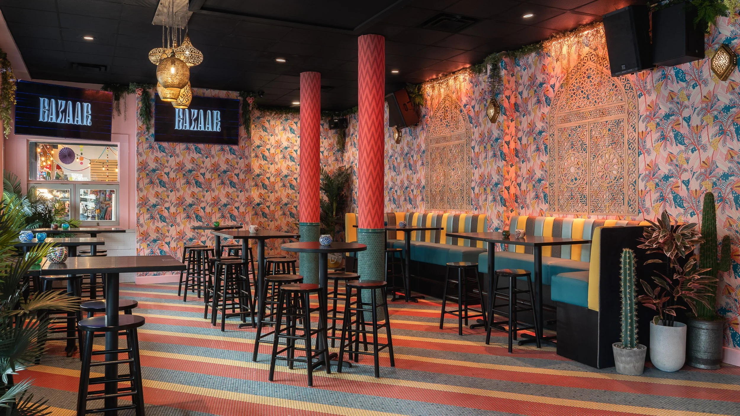 Interior of a vibrant cafe or restaurant with tropical-themed wallpaper, pink and red patterned columns, hanging lanterns, and a long colorful cushioned seating area.