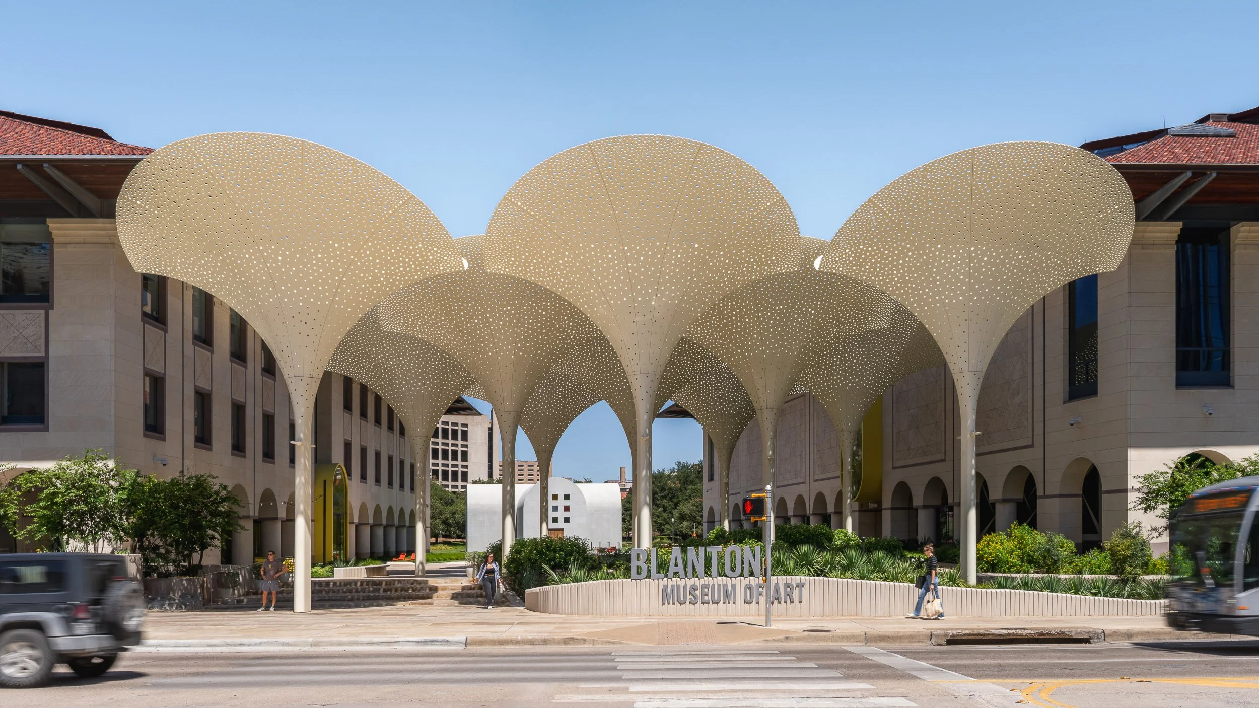 Exterior view of the Blanton Museum of Art in Austin, Texas, featuring a modern architectural canopy with perforated, cloud-like shapes covering a walkway, trees, and pedestrians in the foreground.