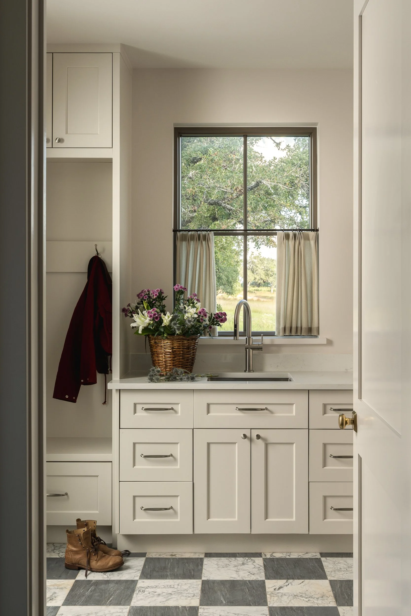 A kitchen with white cabinets, a window with cream curtains, a sink with a bouquet of pink and white flowers, a basket, and a pair of brown boots on a black and white checkered marble floor.