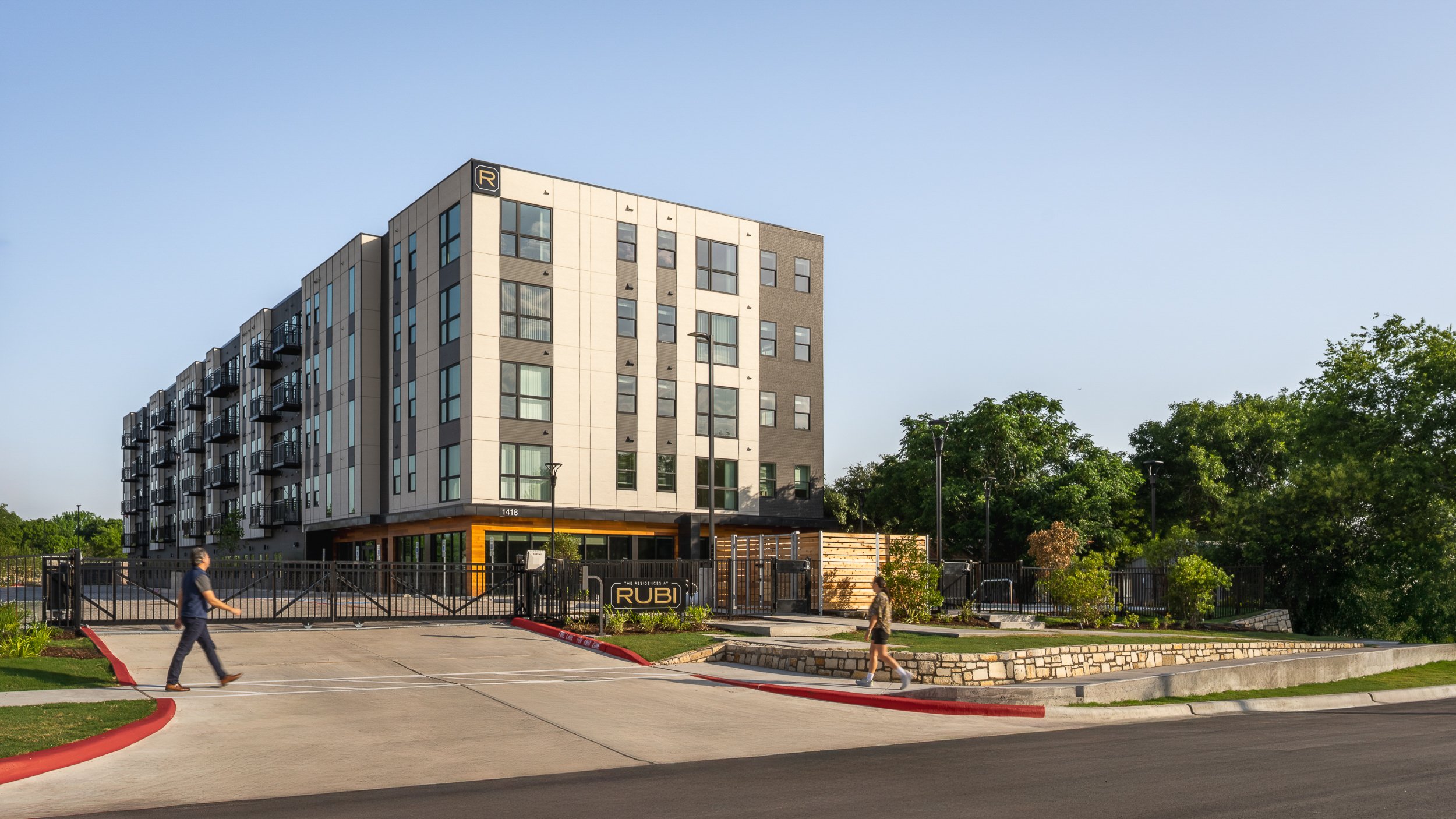 Modern multi-story apartment building with balconies, surrounded by trees, and two pedestrians walking in the foreground. The entrance is gated with a sign that reads 'The Residences at Rubi.'