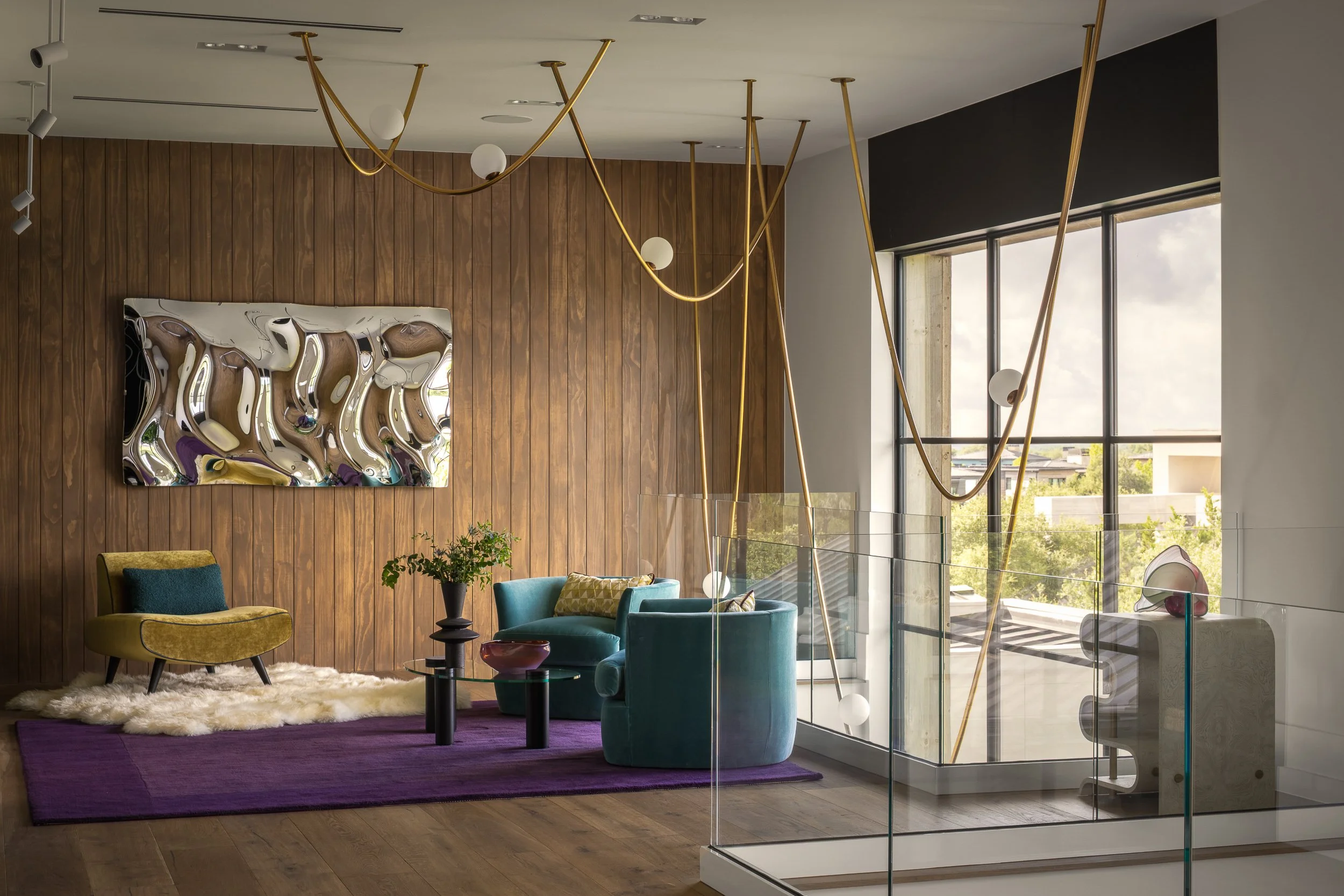 Modern interior living room with wood-paneled wall, colorful armchairs, a textured rug, and a large abstract mirror. A distinctive gold and white lighting fixture hangs from the ceiling near tall windows showing a cityscape and blue sky.