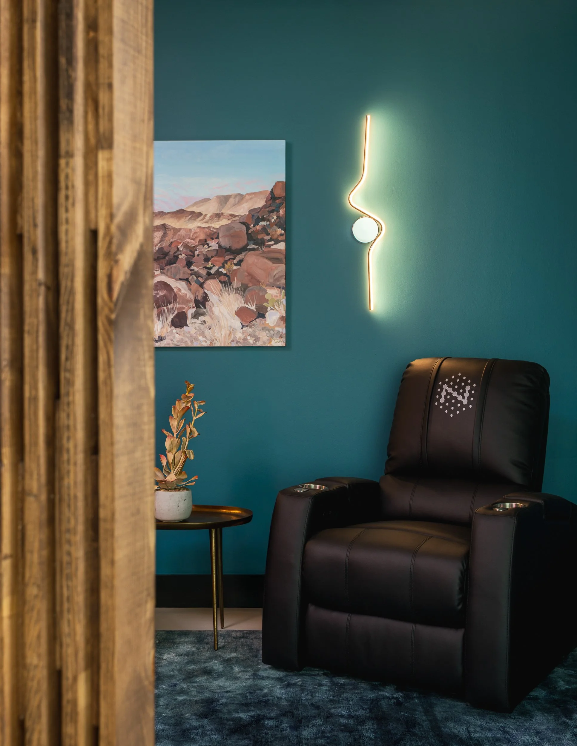 Part of a living room or waiting area, featuring a blue wall with abstract landscape art, a modern neon wall sculpture, a black leather armchair, a side table with a potted plant, and a wooden room divider.