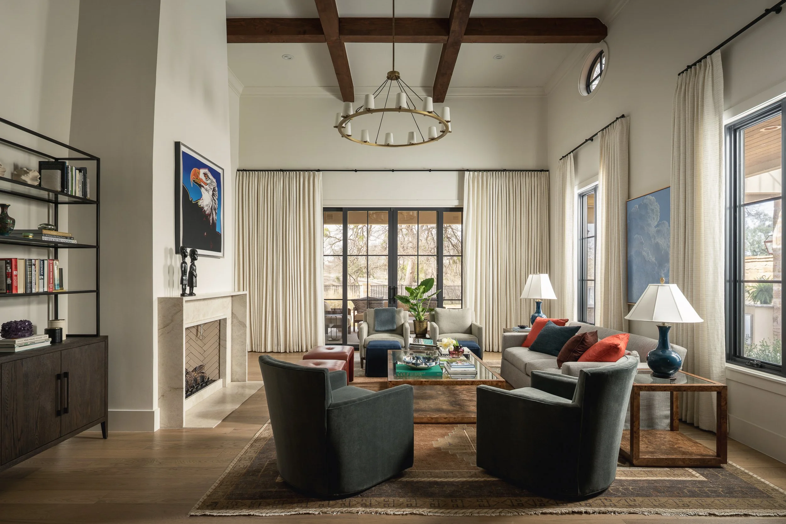 A spacious living room with high ceilings and exposed wooden beams. There are large windows with white curtains, a fireplace with decorative statues, a bookshelf, and various chairs and sofas, along with lamps and plants.