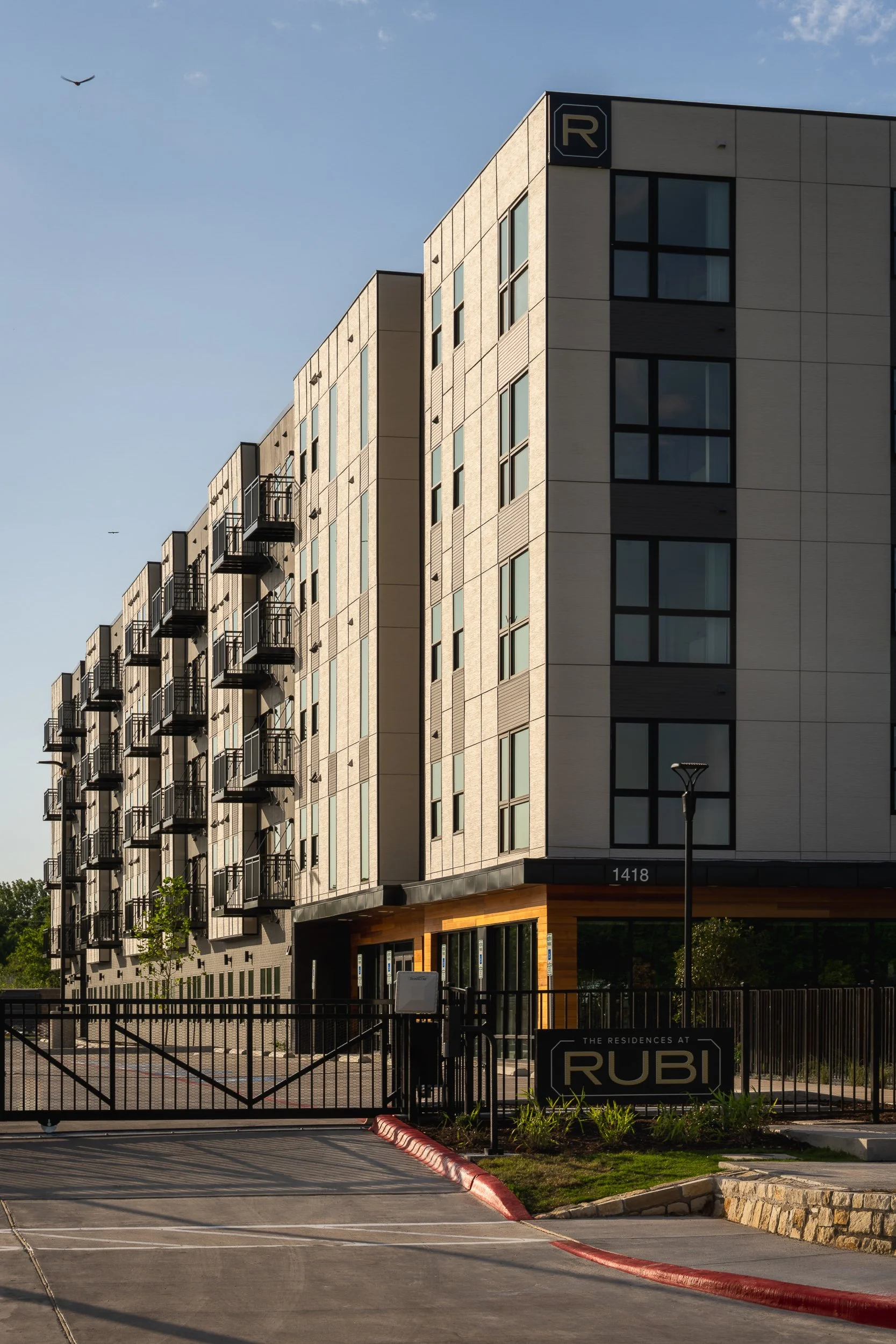 Modern multi-story residential building with a sign reading 'The Residences at Rubi' and a gated entrance in the foreground.