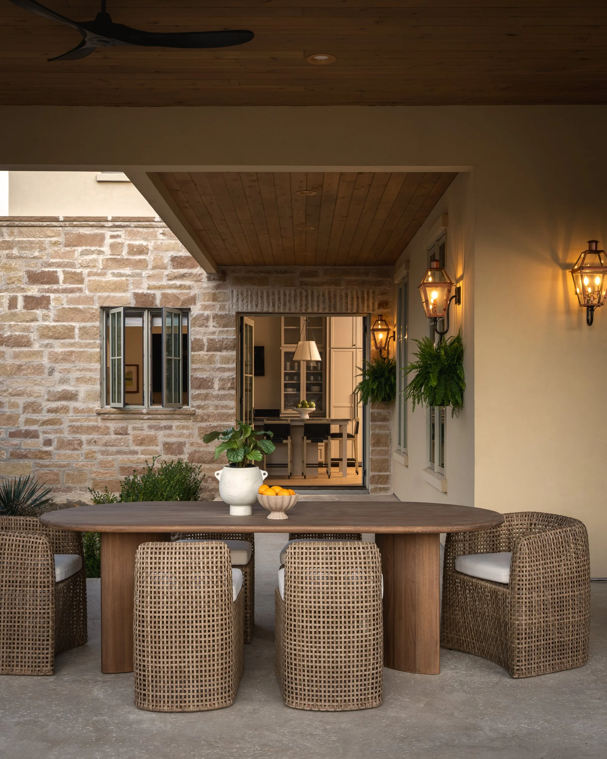 Outdoor patio with tan wicker chairs around a wooden table decorated with a white vase of green plants and a bowl of oranges. The background features a brick wall, open French doors leading inside, and wall-mounted lanterns with warm lighting. The ce