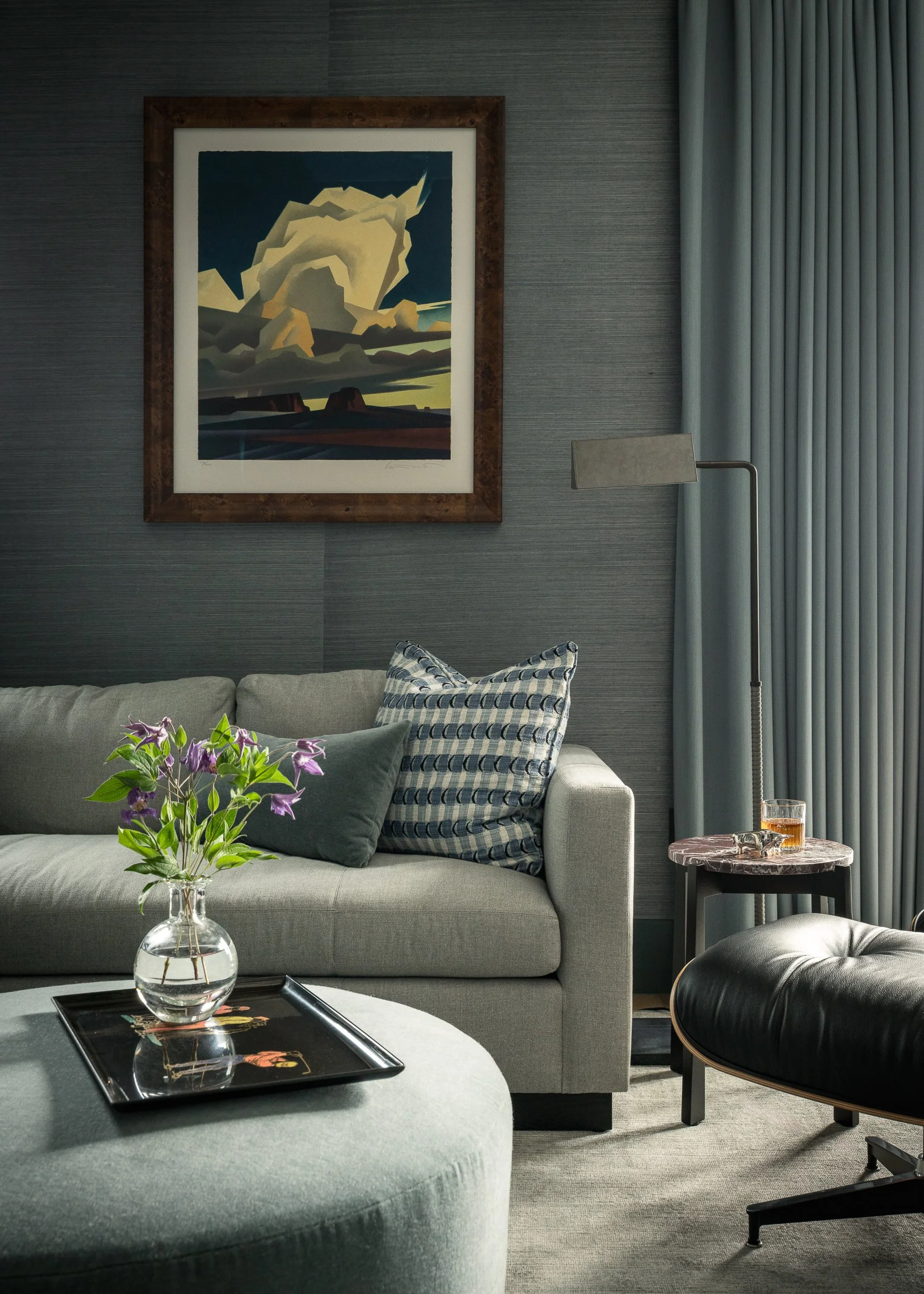 Living room with a beige sofa, decorative pillows, a glass vase with purple flowers on a coffee table, a modern black armchair, a floor lamp, a framed landscape picture on the wall, and curtains by the window.