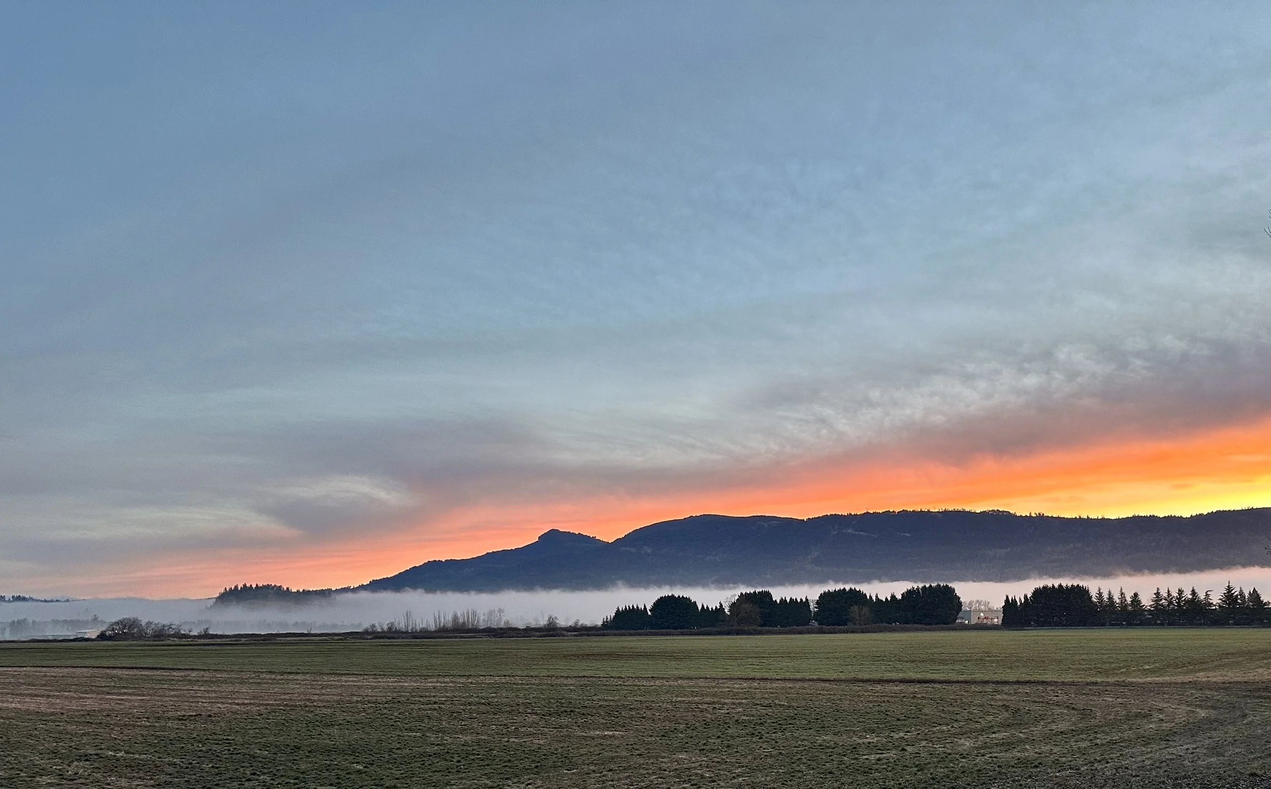 A sunset over a mountain range with a colorful sky transitioning from pink and orange near the horizon to blue higher up, and a foggy landscape with fields and trees in the foreground.