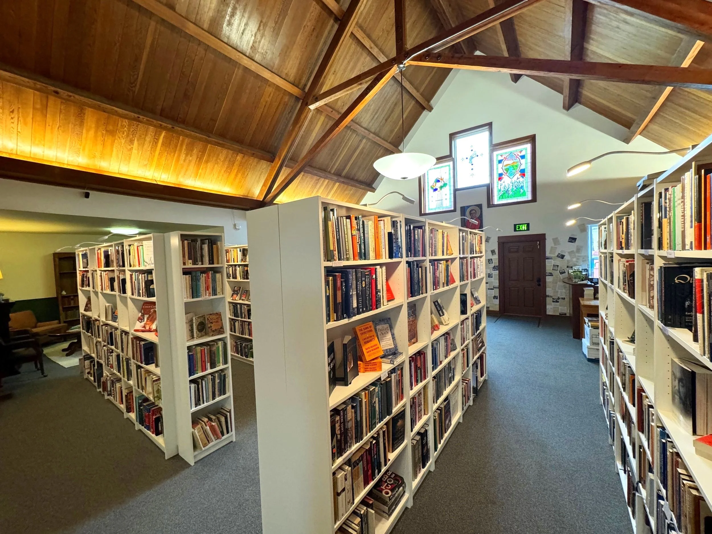 Interior of a bookstore or library with white bookshelves filled with books. The ceiling is high with wood paneling and exposed beams, and there are stained glass windows on the wall letting in natural light. A sign with green lights shows an exit door at the back.