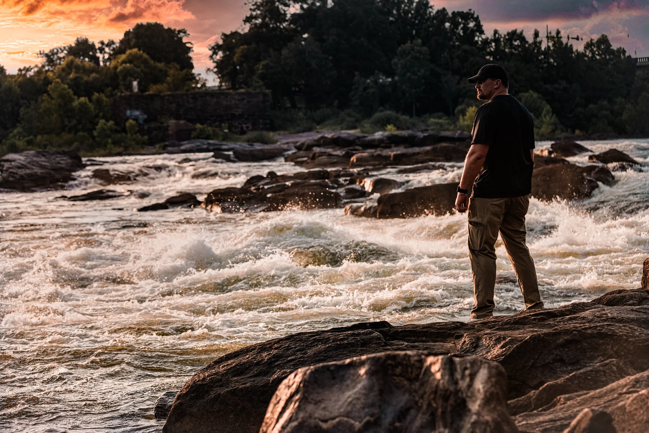 Environmental portrait of a man standing on rocks beside a flowing river at sunset.